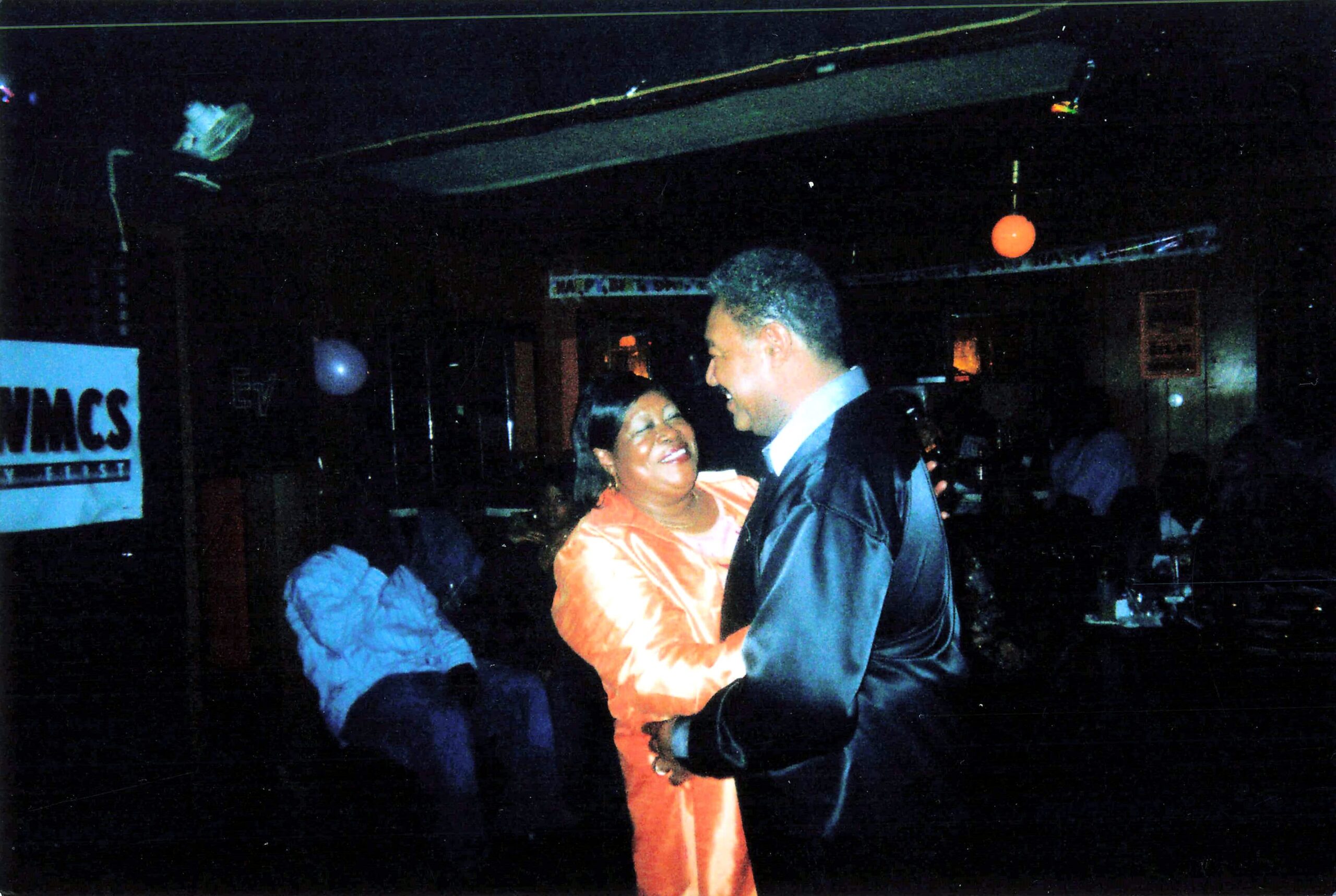 Couple dancing closely at a dim party venue; woman in an orange dress smiles at the man in a dark suit under party lighting with an orange balloon above them