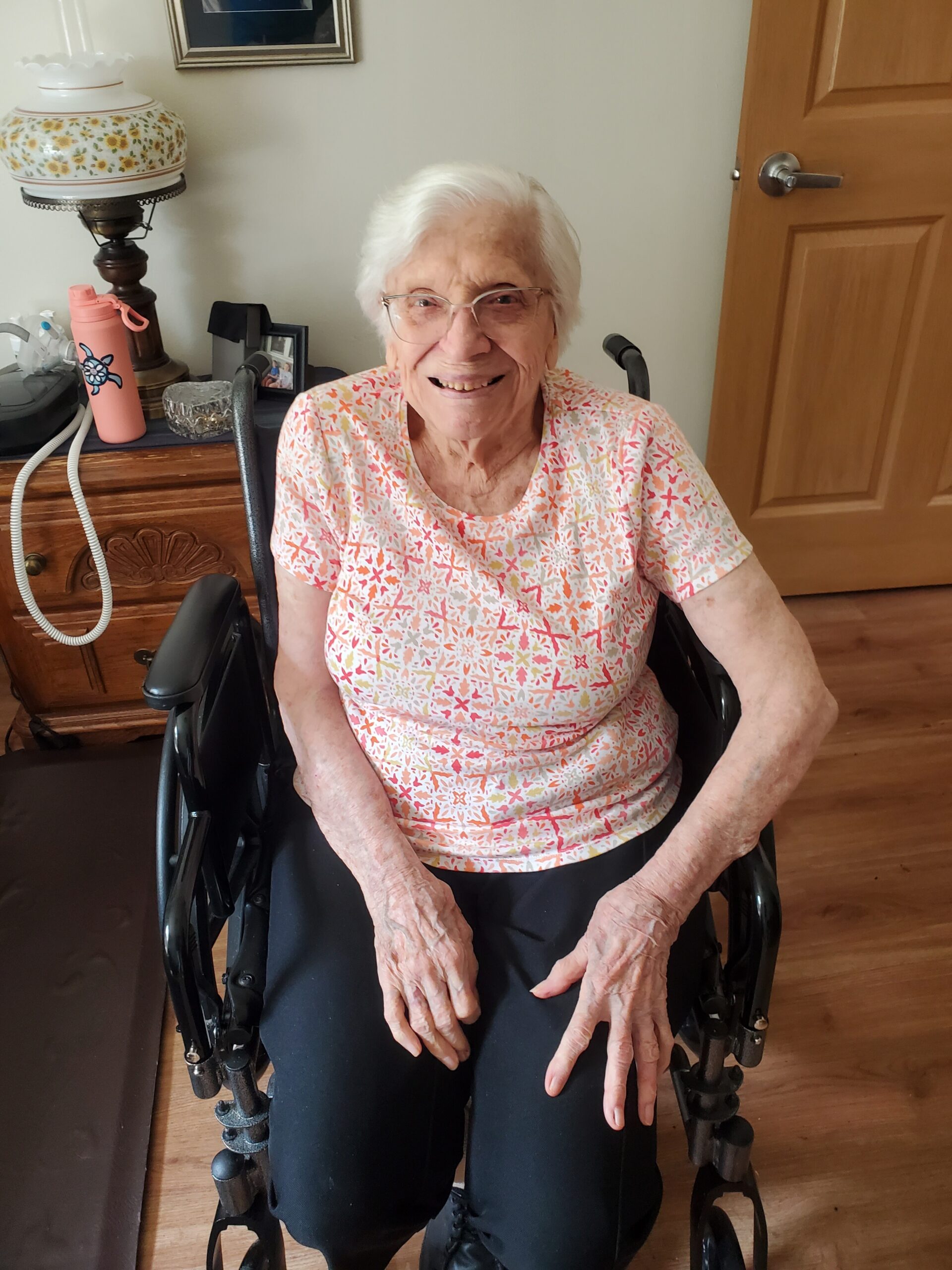 Smiling elderly woman in a wheelchair indoors, wearing a pink patterned blouse and black pants, hands resting on her lap on a wooden floor.