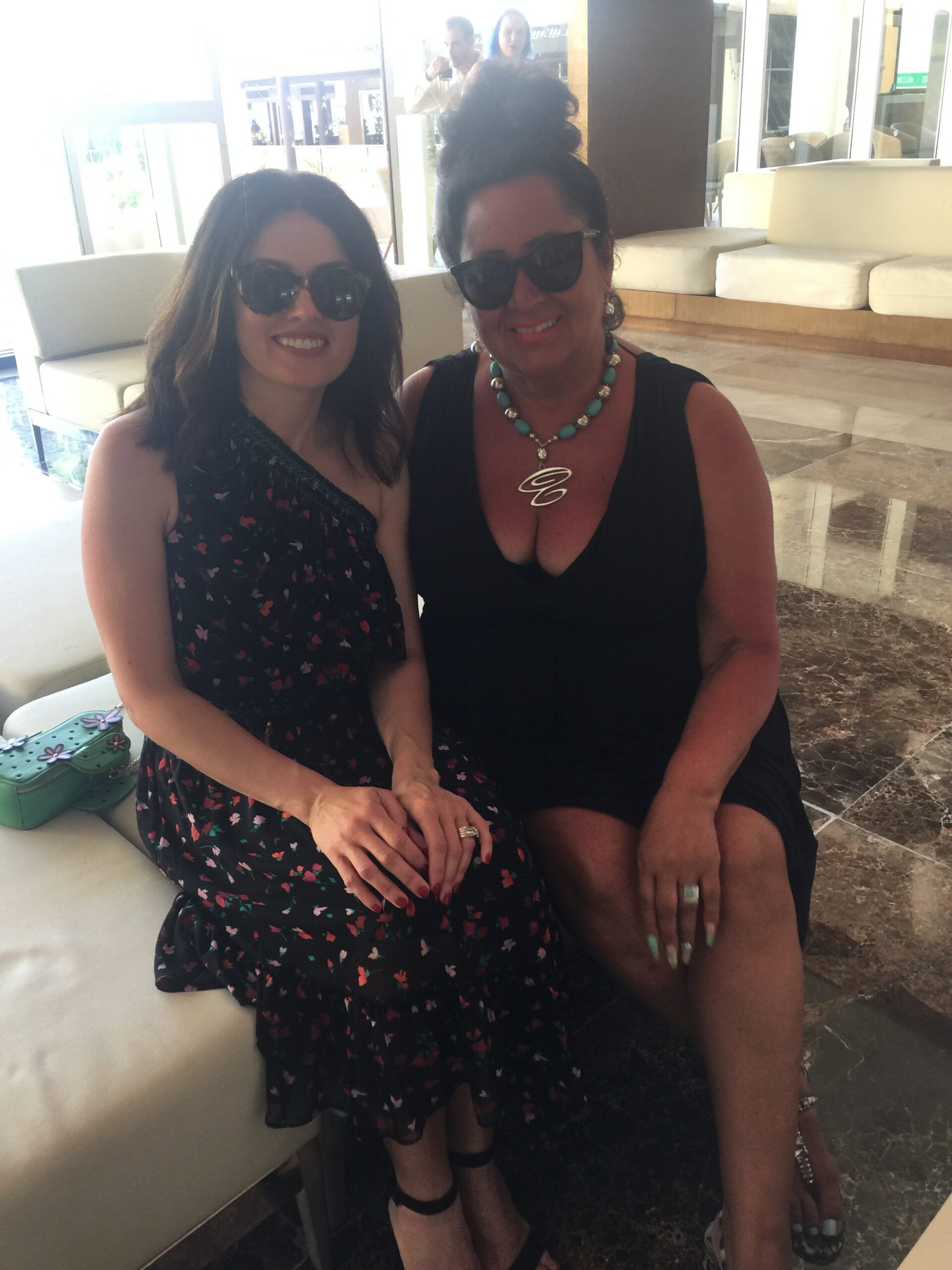 Two women wearing sunglasses sit closely on a white couch in a bright lobby, smiling at the camera.