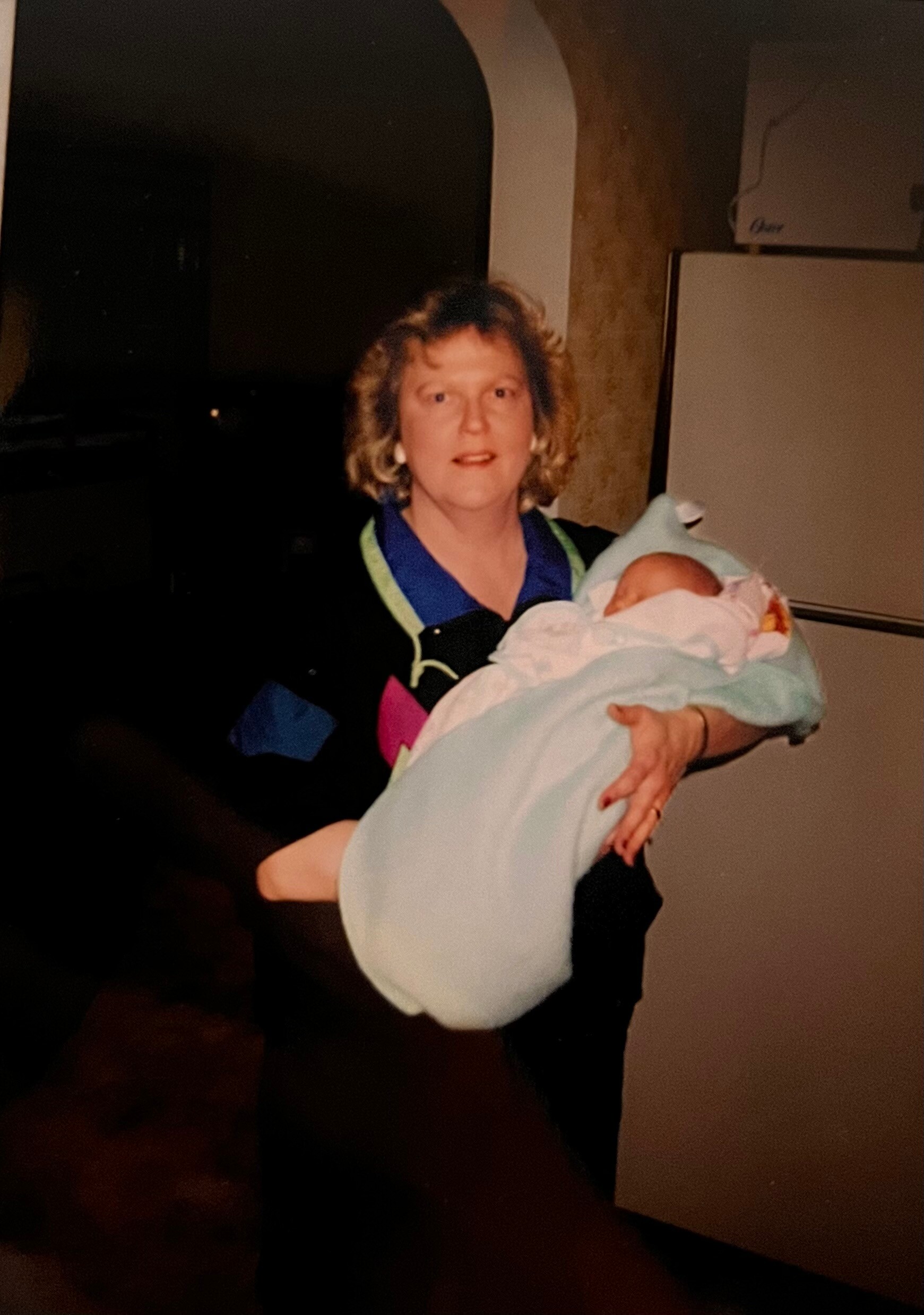 Woman holding a newborn baby wrapped in a light blue blanket indoors, facing camera.