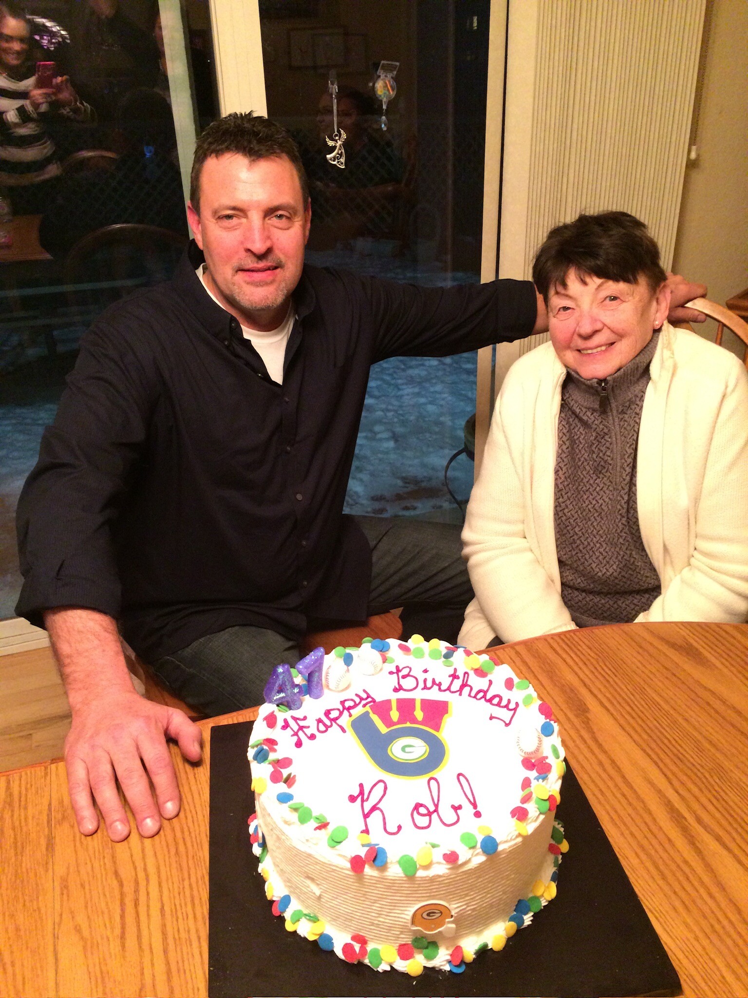 Man and an elderly woman sit together at a table with a colorful birthday cake that reads 'Happy Birthday' in red letters.