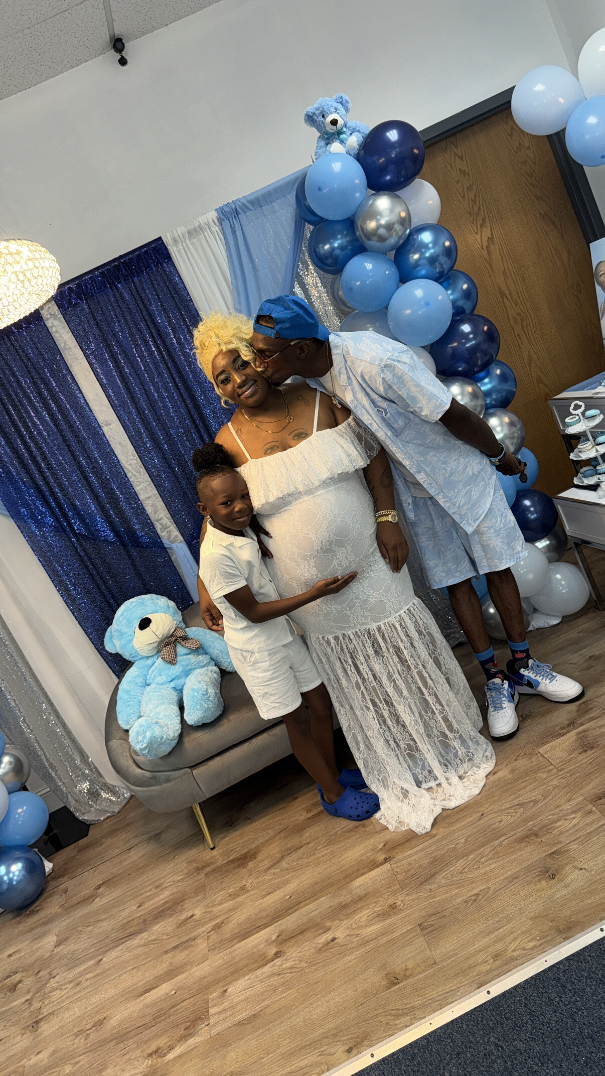 Pregnant woman in a white lace dress stands with a man kissing her on the shoulder and a young girl hugging her, at a blue‑themed party with balloons and a teddy bear backdrop.