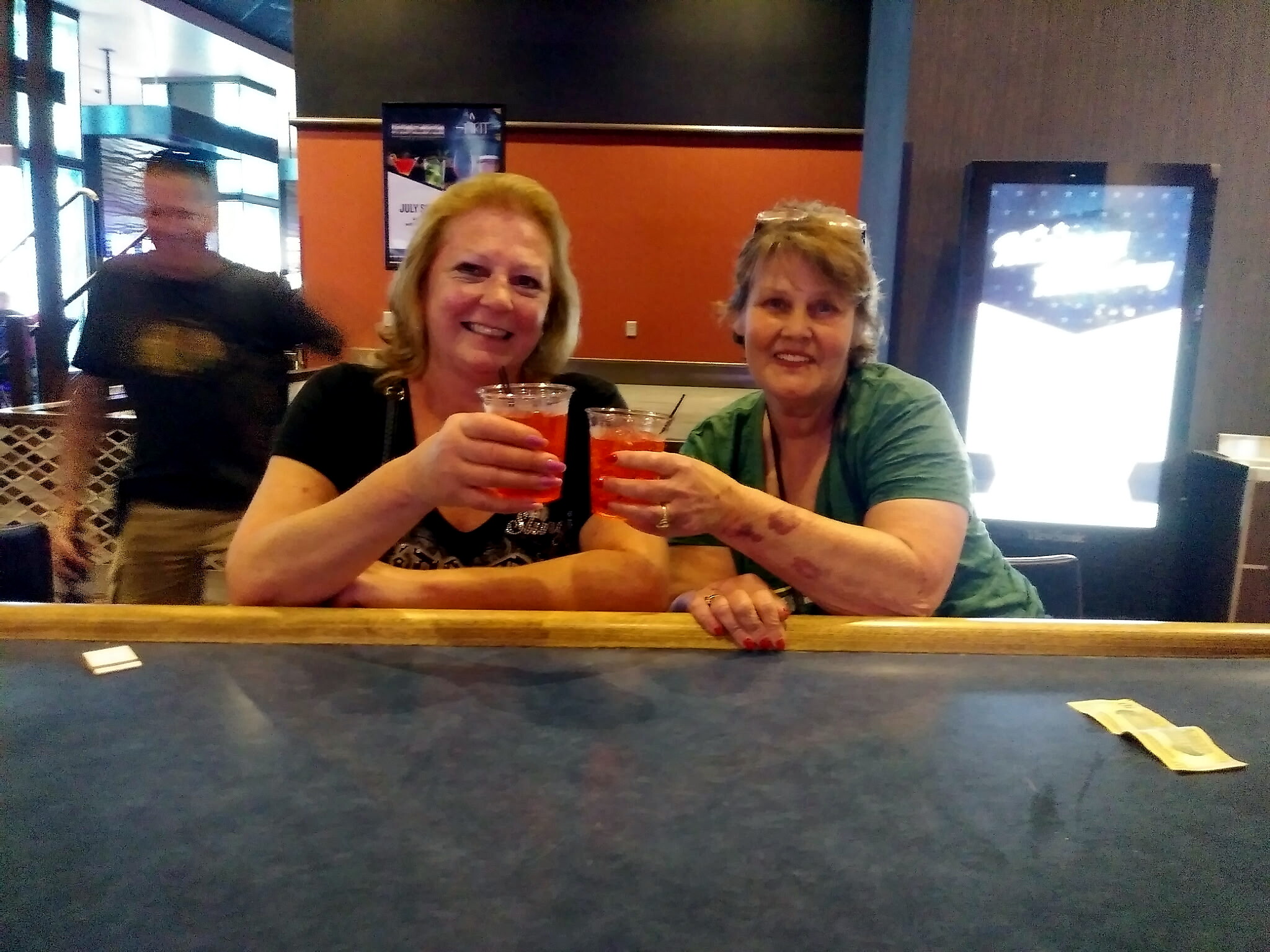 Two smiling women sit at a bar toasting with red/orange cocktails. A man walks in the background.