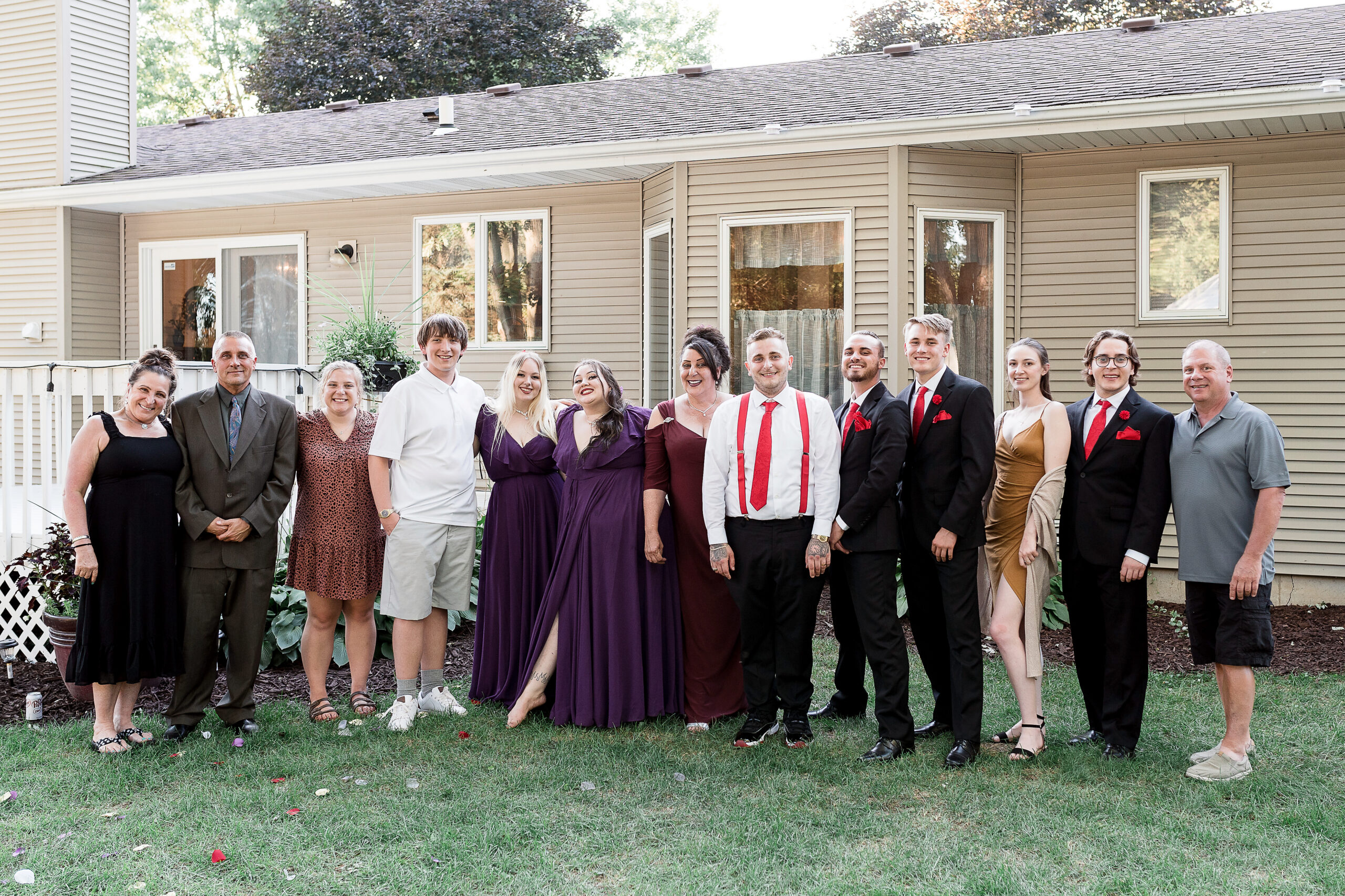 Group of people in formal attire posing on a lawn in front of a beige house, wearing gowns and suits, smiling for a photo.