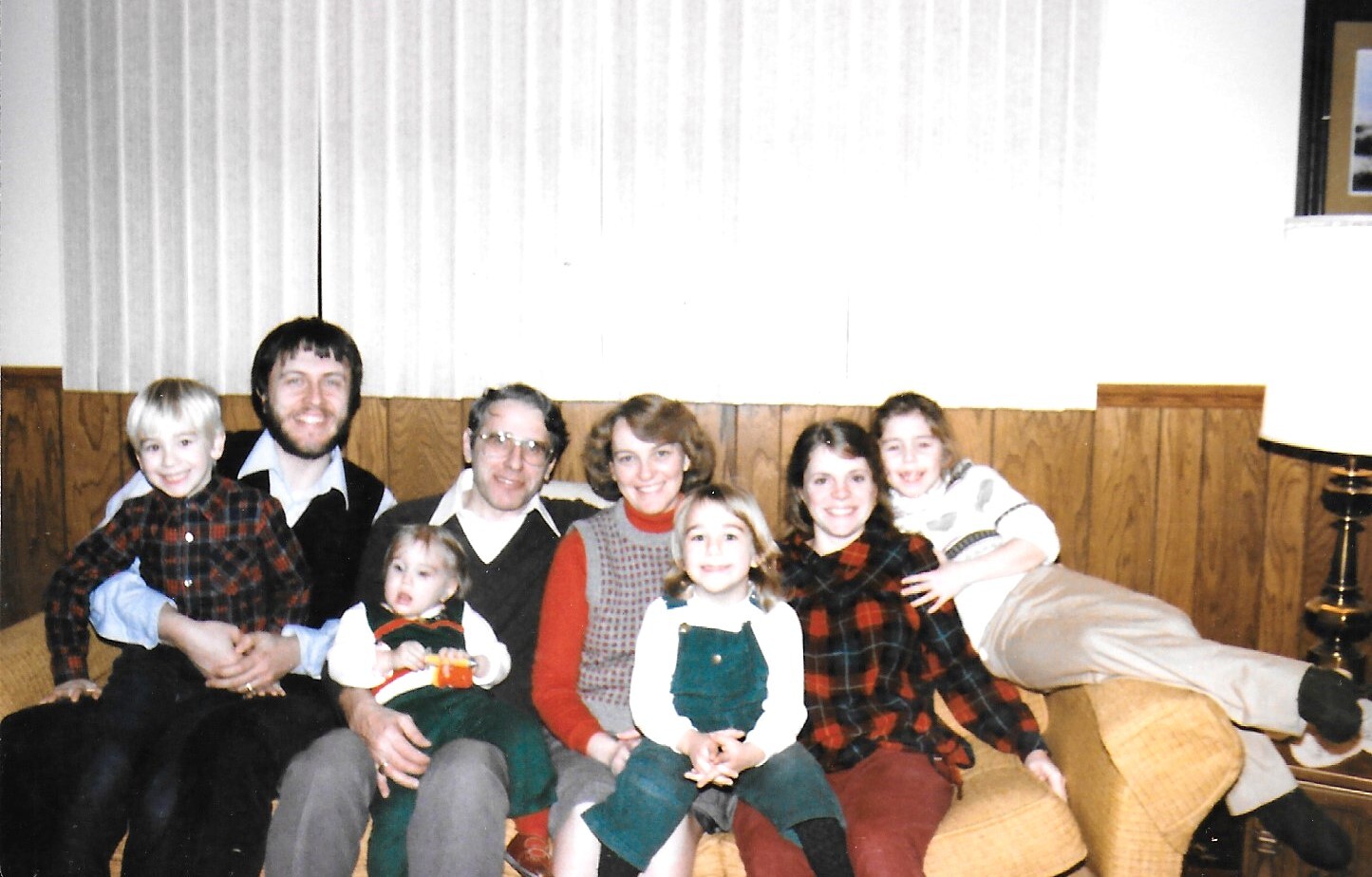 A multi-generational family sits together on a tan sofa in a retro living room, smiling at the camera; adults and children wearing plaid and knit outfits from the 1970s.