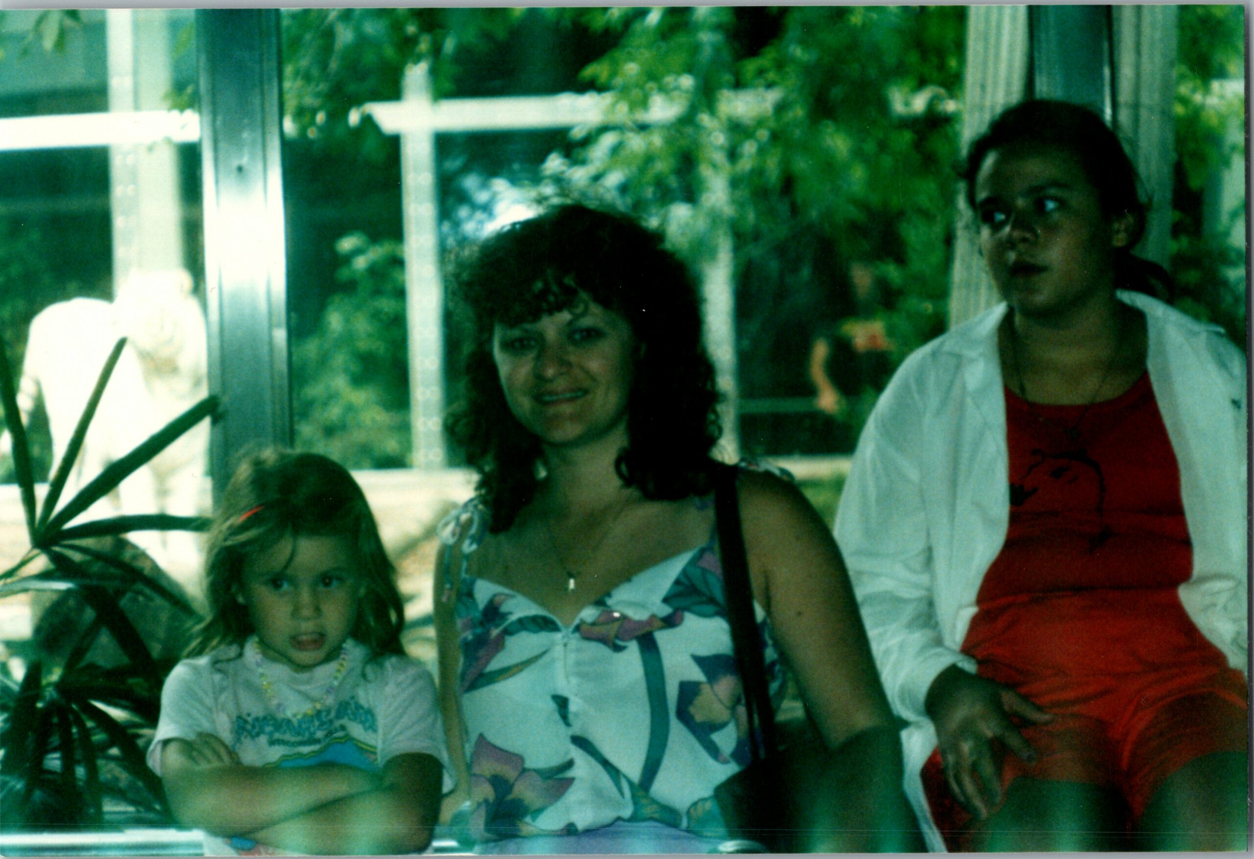 Three people posing together indoors beside a window, with a woman in a patterned blouse center and a girl on the left wearing a white shirt, and a woman in red on the right.
