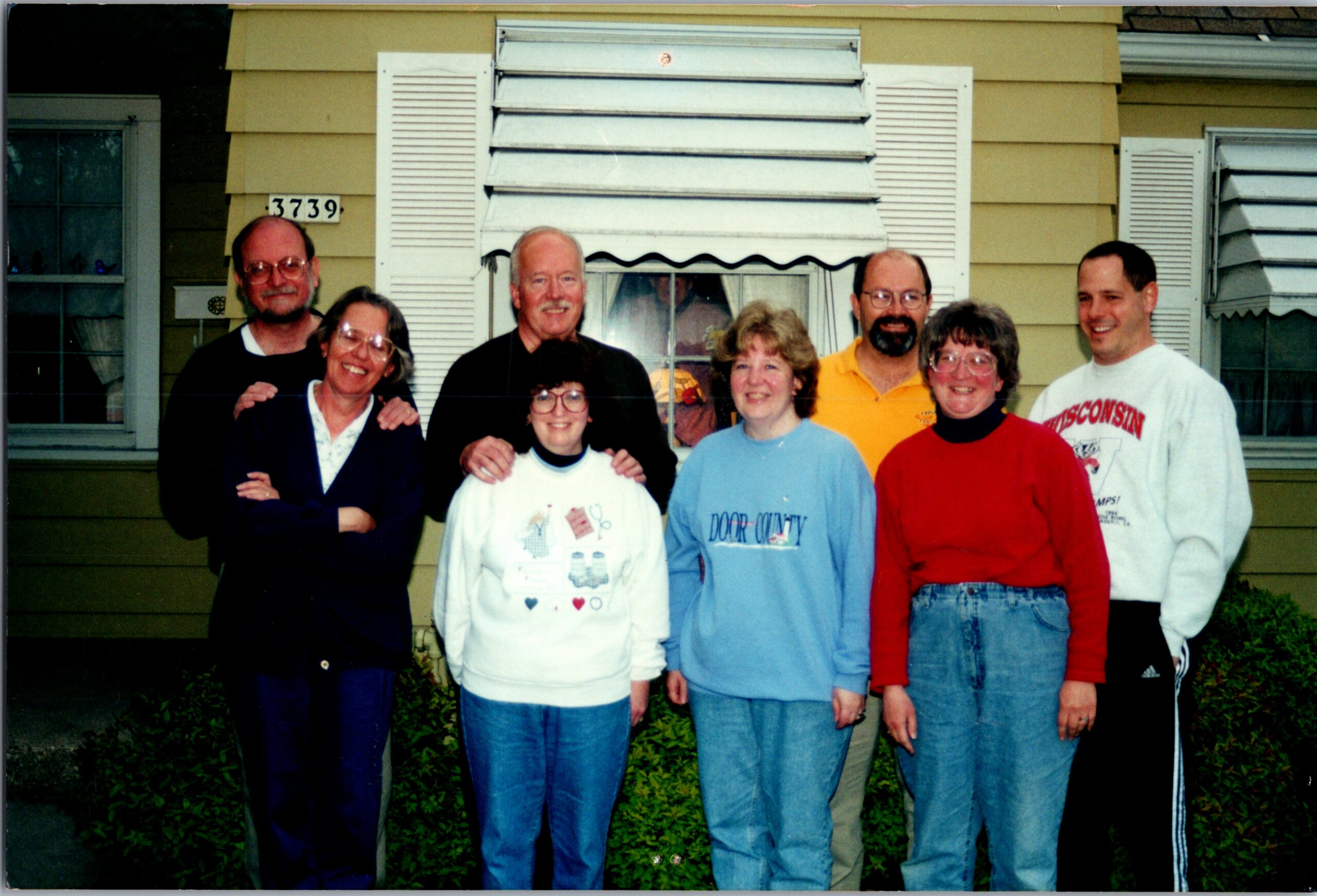 Seven adults posing for a casual group photo in front of a yellow house with white shutters (house number 3739) and greenery.
