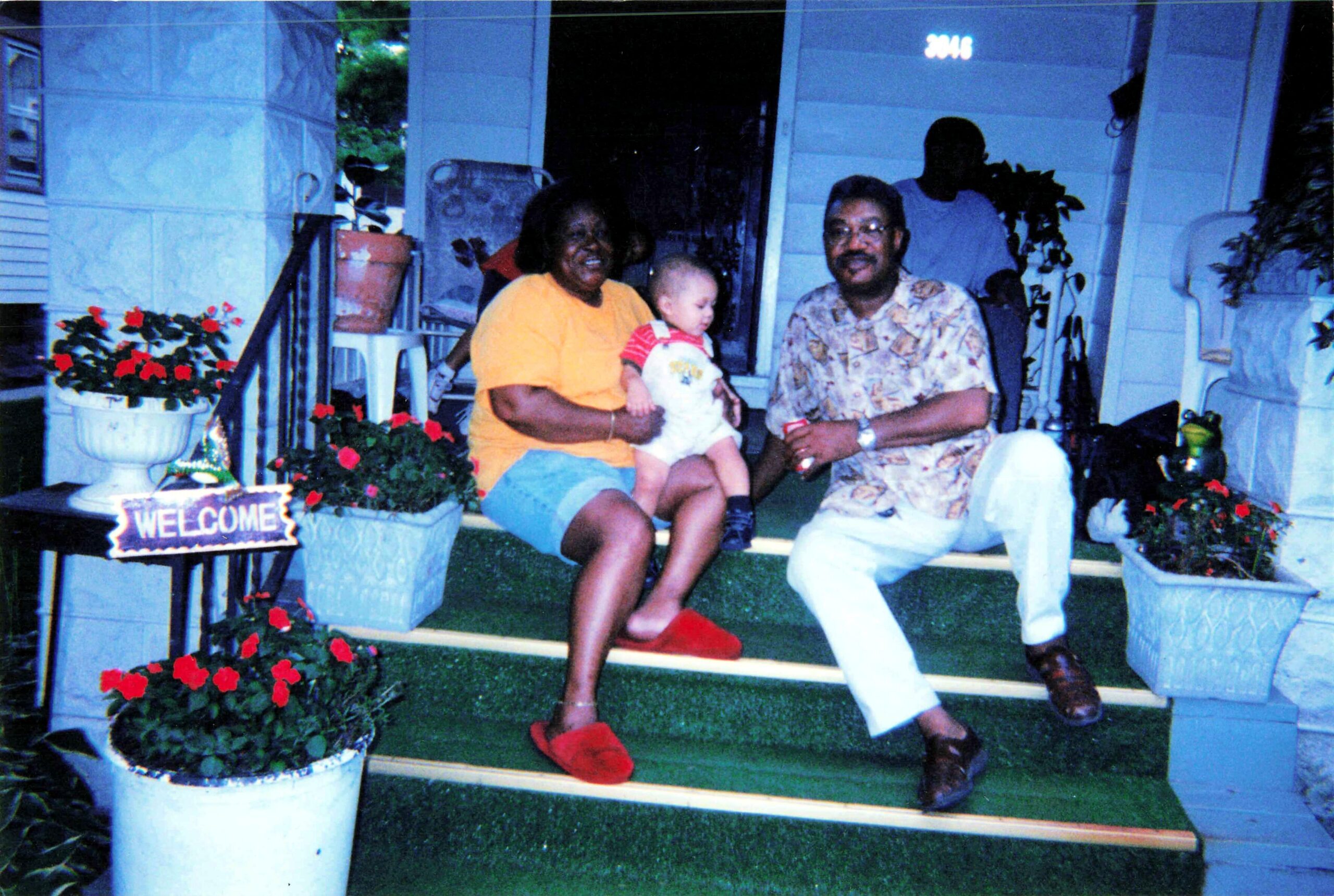 A woman in a yellow shirt holding a baby sits on porch steps next to a man in a patterned shirt; red flowers in planters and a 'Welcome' sign nearby.