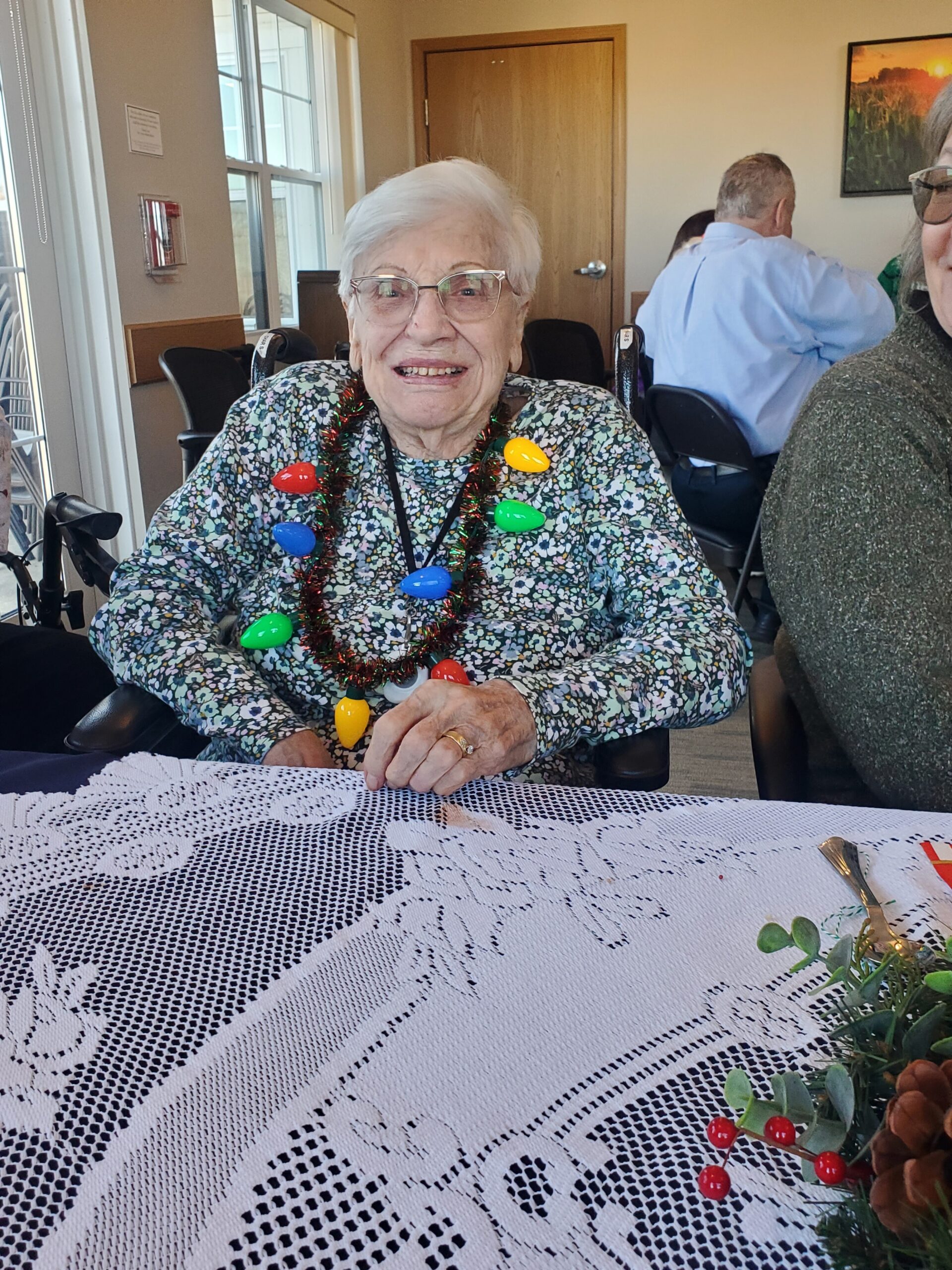 Smiling elderly woman in glasses wearing a floral blouse with colorful holiday light necklace, seated at a lace-covered table in a gathering venue.