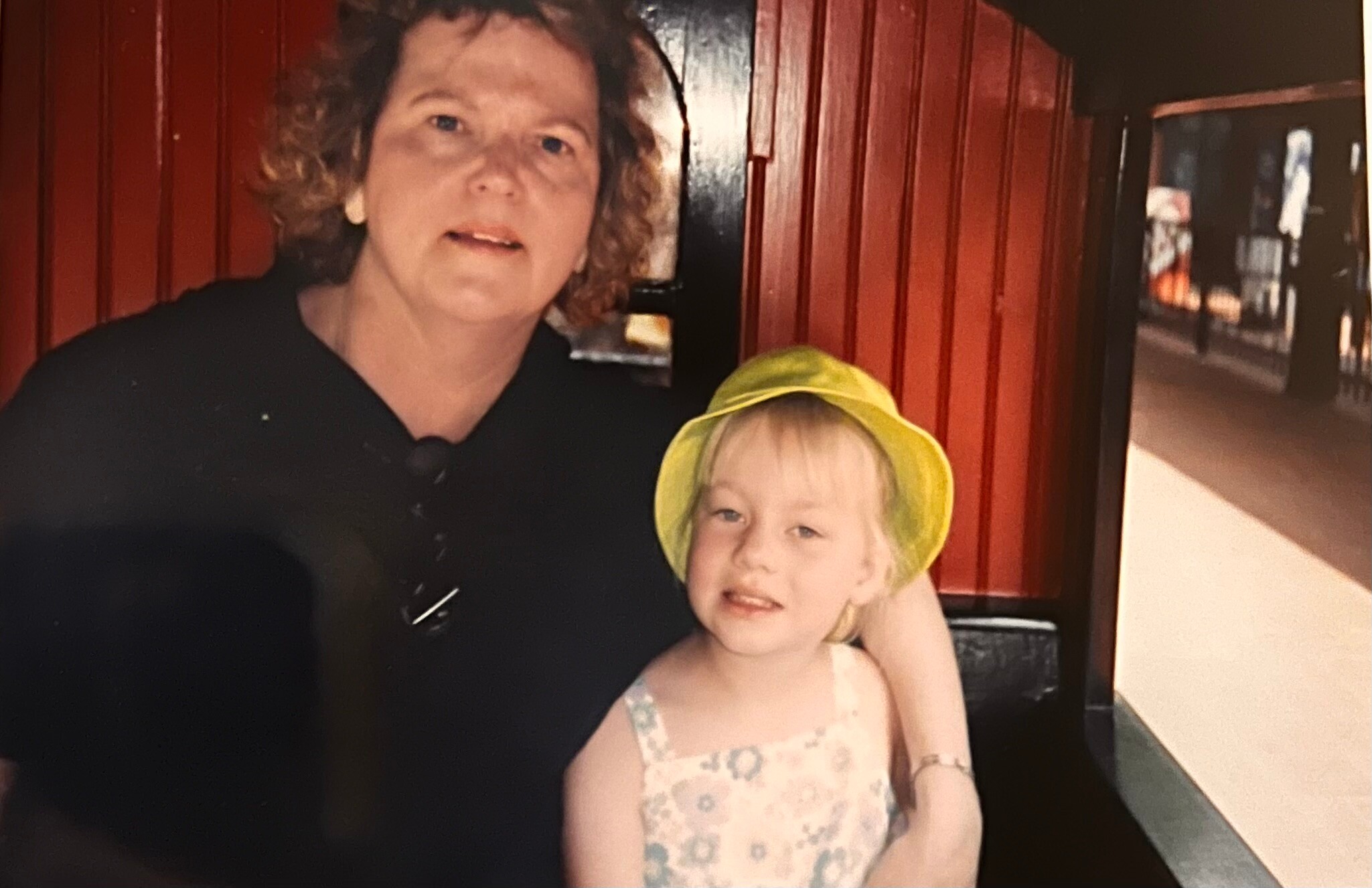 Two people pose for a photo indoors: an adult woman with short curly hair beside a young girl wearing a bright yellow hat, against reddish wooden paneling behind them.