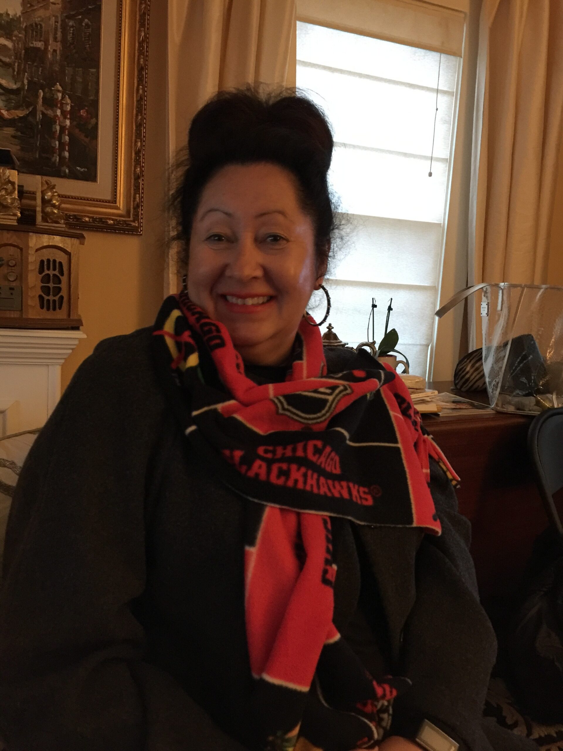 Smiling woman indoors wearing a red and black Chicago Blackhawks scarf, with curtains and a framed painting in the background.