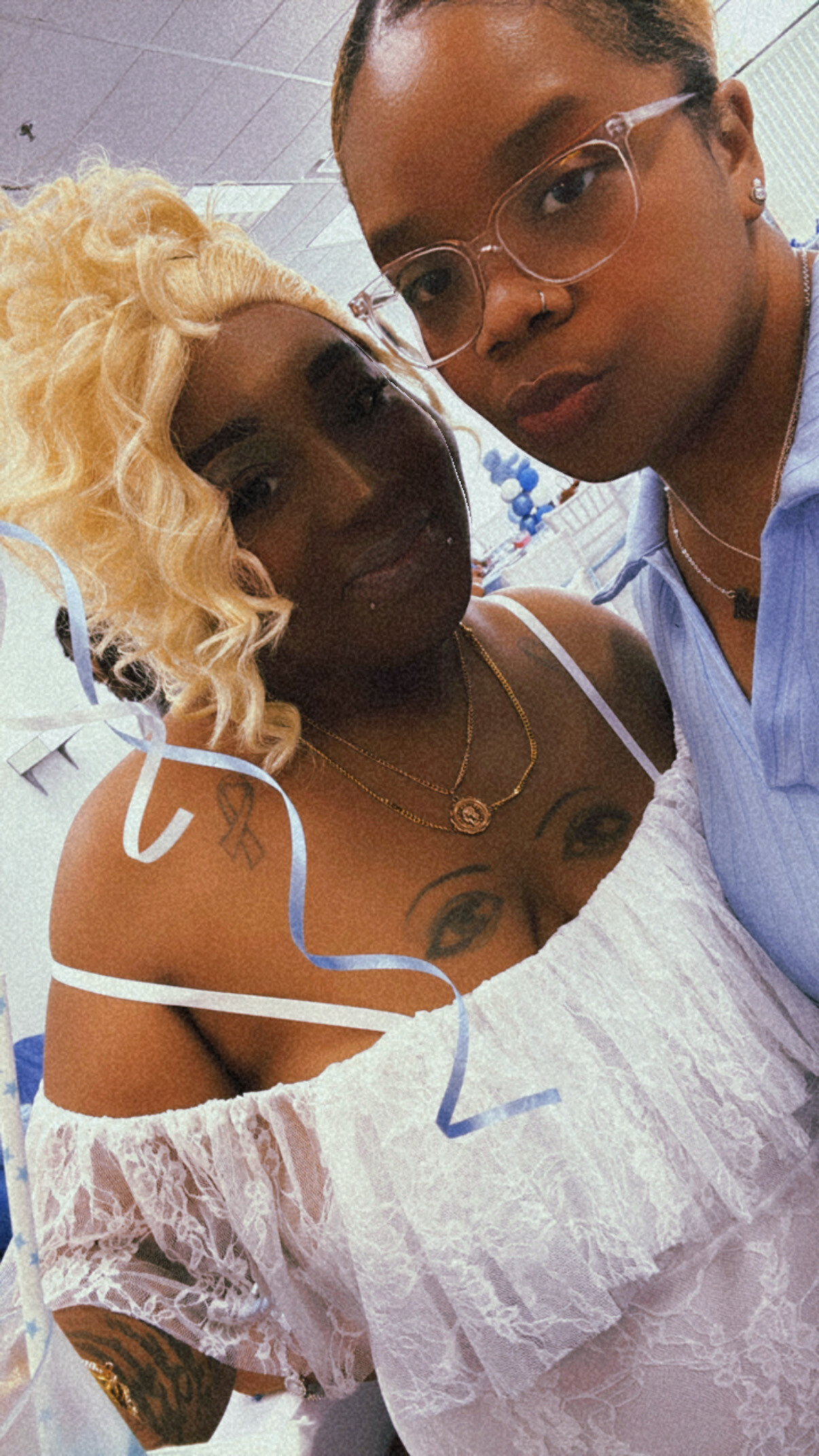 Two women pose for a selfie close up; one with blonde curls in a lace top, the other with clear glasses and a nose ring.