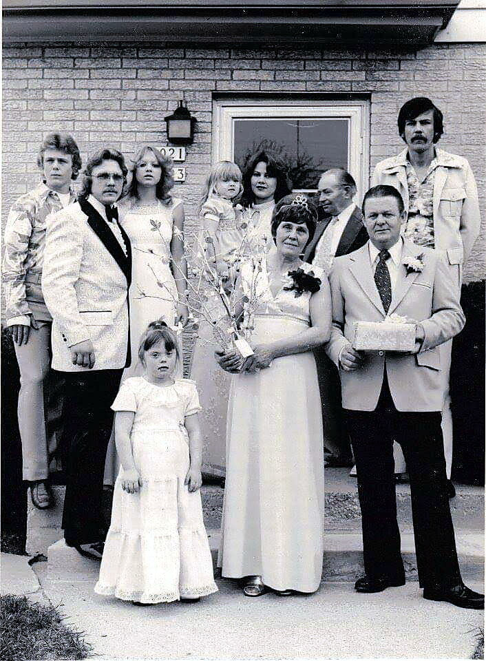 Wedding party posing on a brick house porch: bride in a white gown holding a bouquet, groom in a light suit, and several guests in 1970s attire including two young girls in dresses.