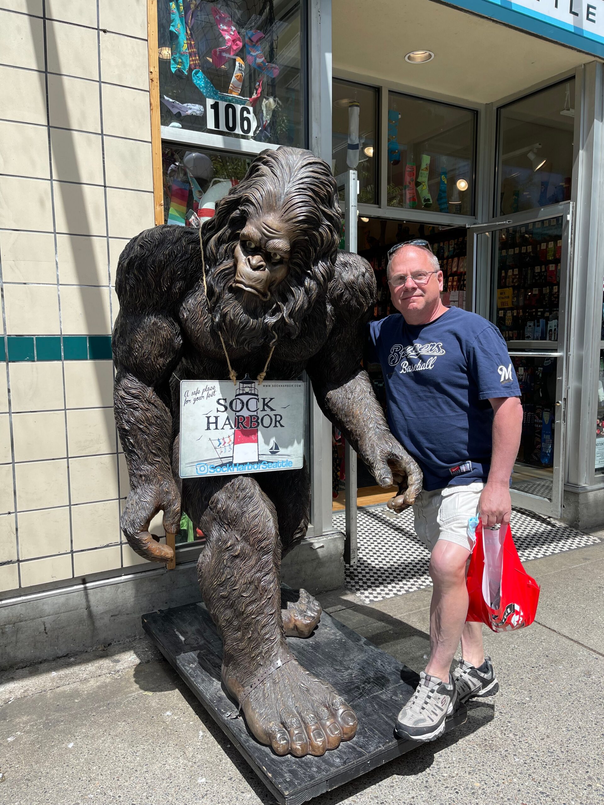 Man in a navy t-shirt posing beside a life-size gorilla statue outside a store, holding a red shopping bag.