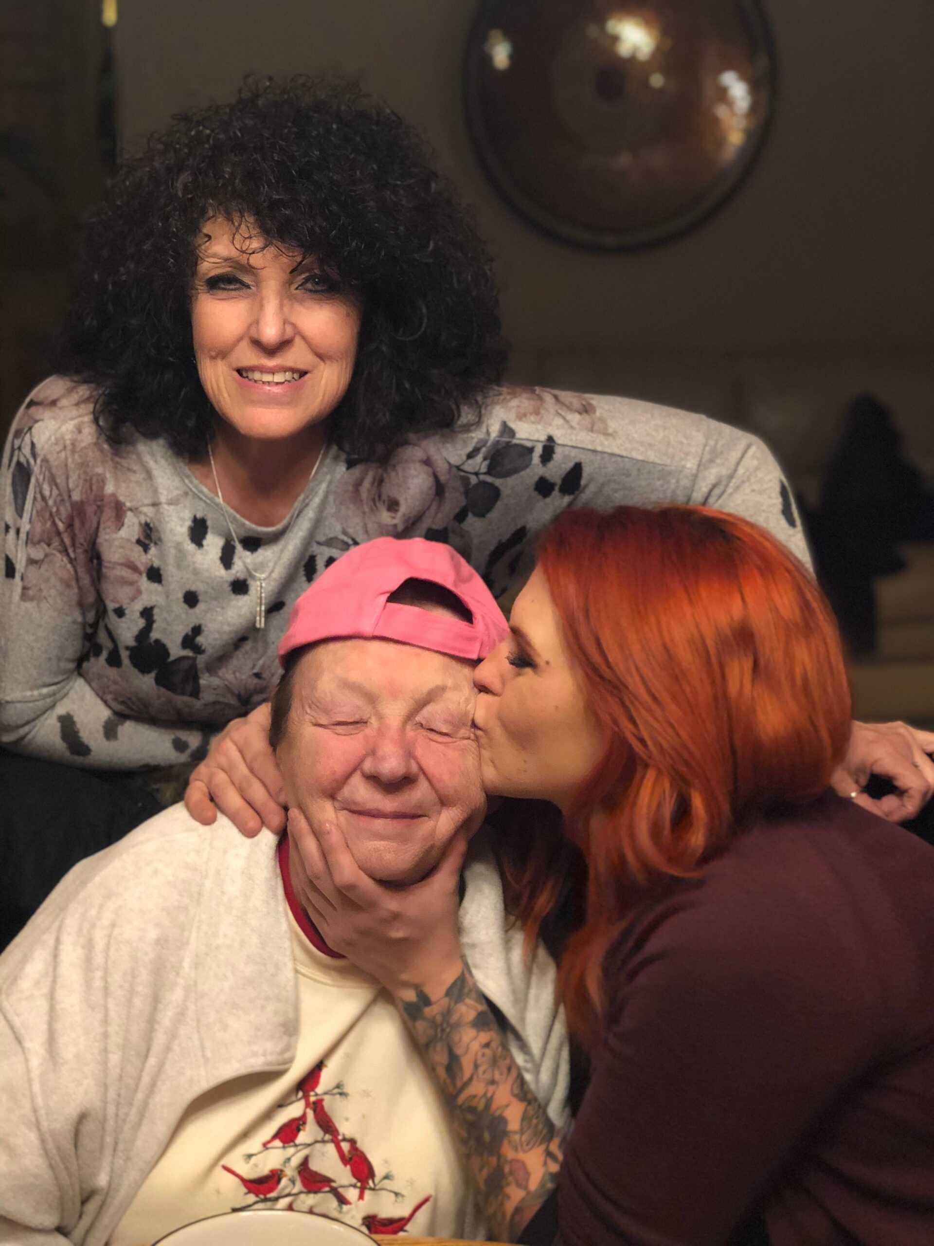 Smiling woman with curly hair leans over an elderly man while a red-haired friend kisses his cheek in a cozy group hug at a table.