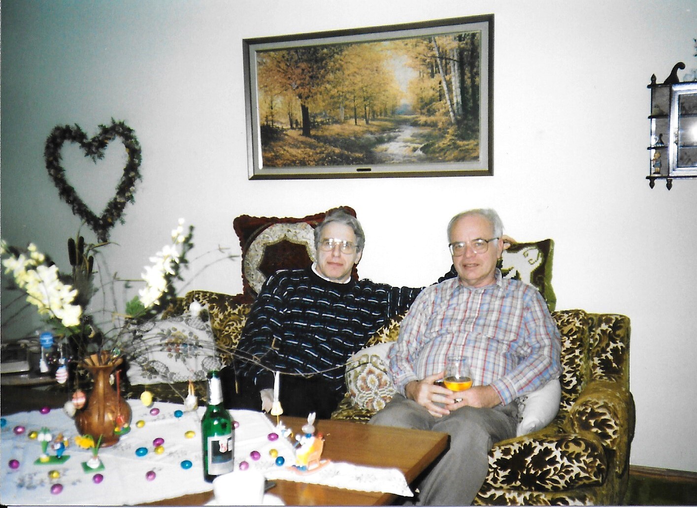Two elderly people sit close together on a patterned sofa in a cozy living room, holding drinks and smiling at the camera; a table with decorations sits in front of them.