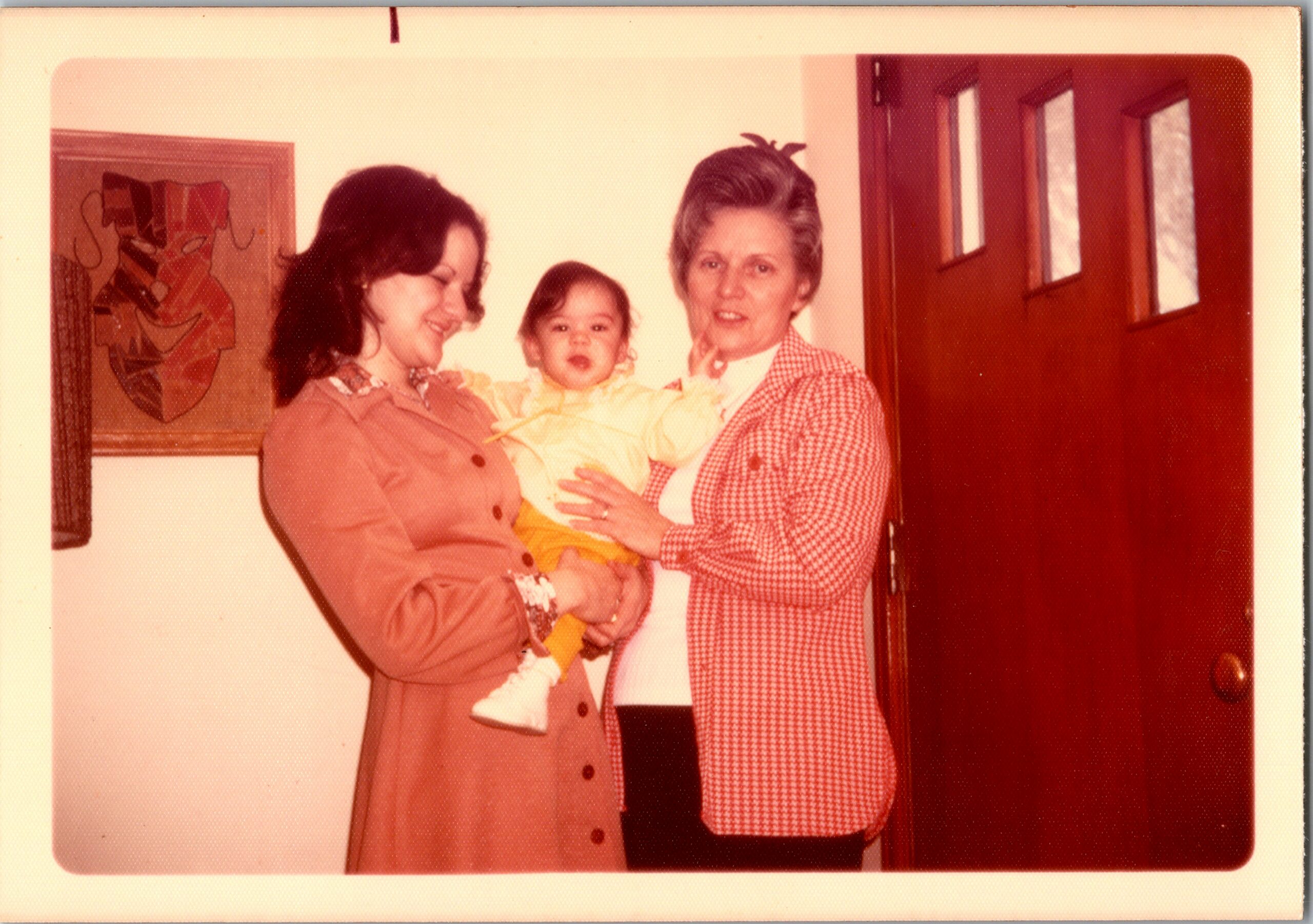 Two women hold a smiling baby in a home hallway.