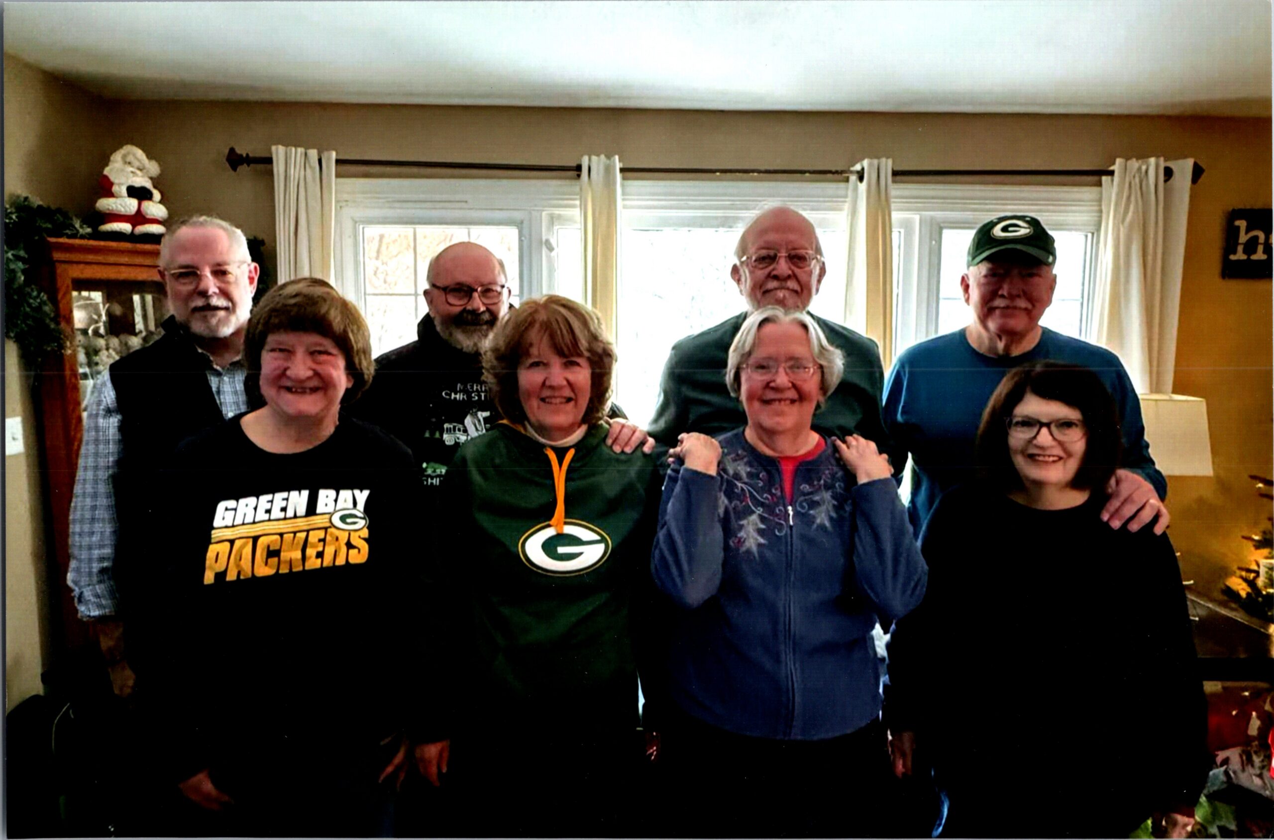 Group of eight adults posing together in a living room, some wearing Green Bay Packers apparel and hats, smiling for a casual photo.