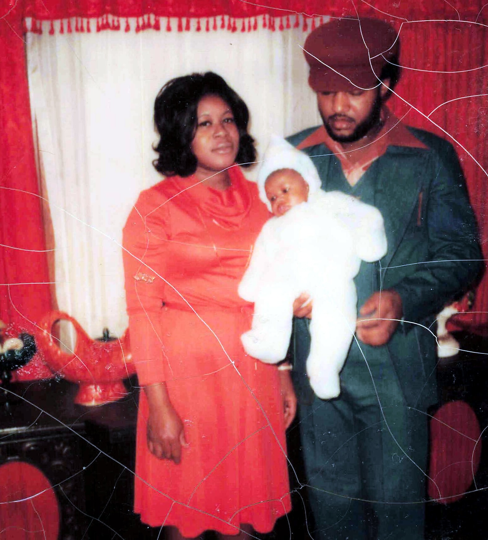 Woman in a red dress holds a swaddled baby in white, standing next to a man in a green suit and brown cap against red curtains.
