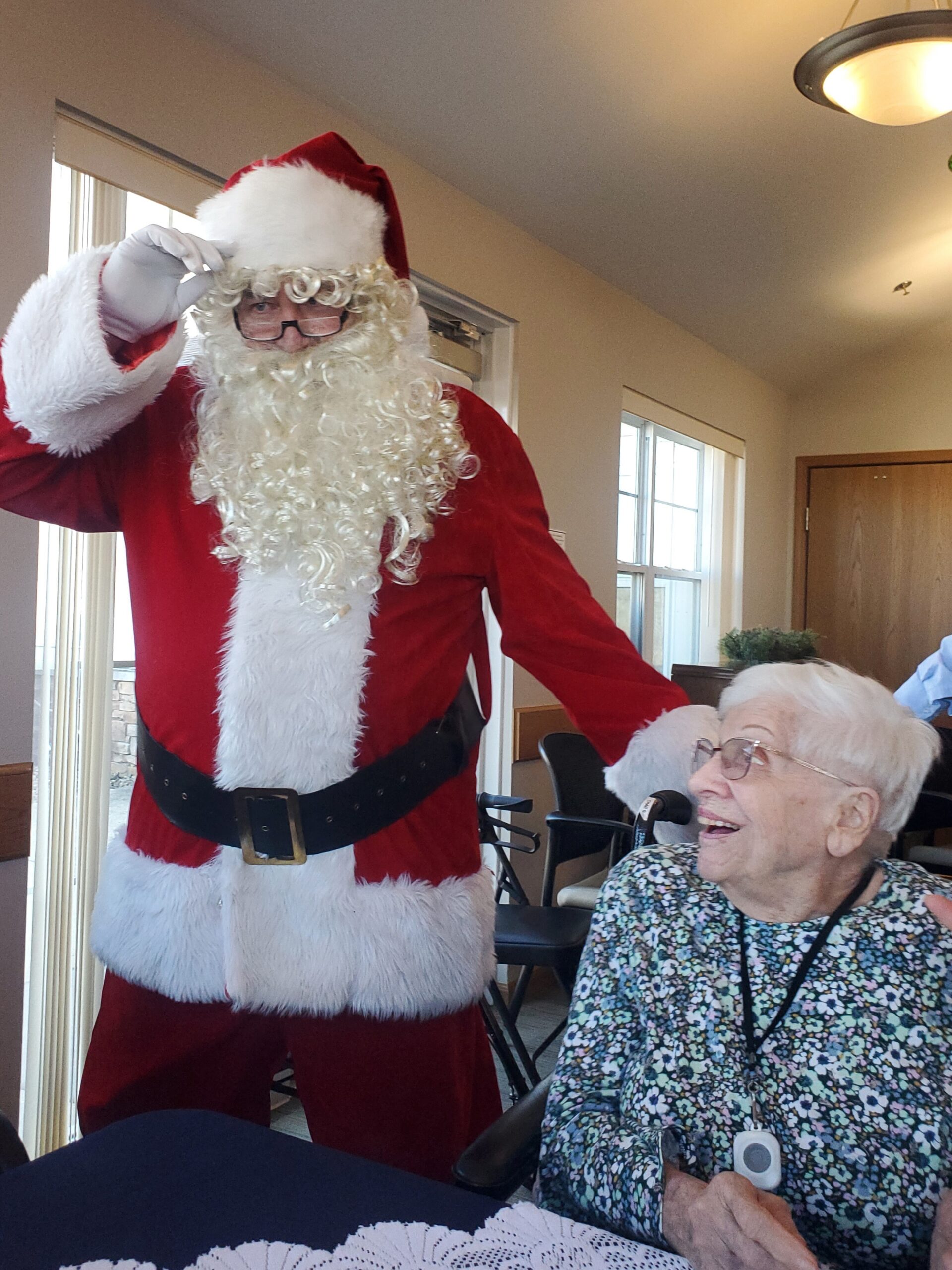 Santa Claus in a red suit and white beard salutes at a holiday visit, standing beside an elderly woman in a wheelchair who smiles warmly indoors.