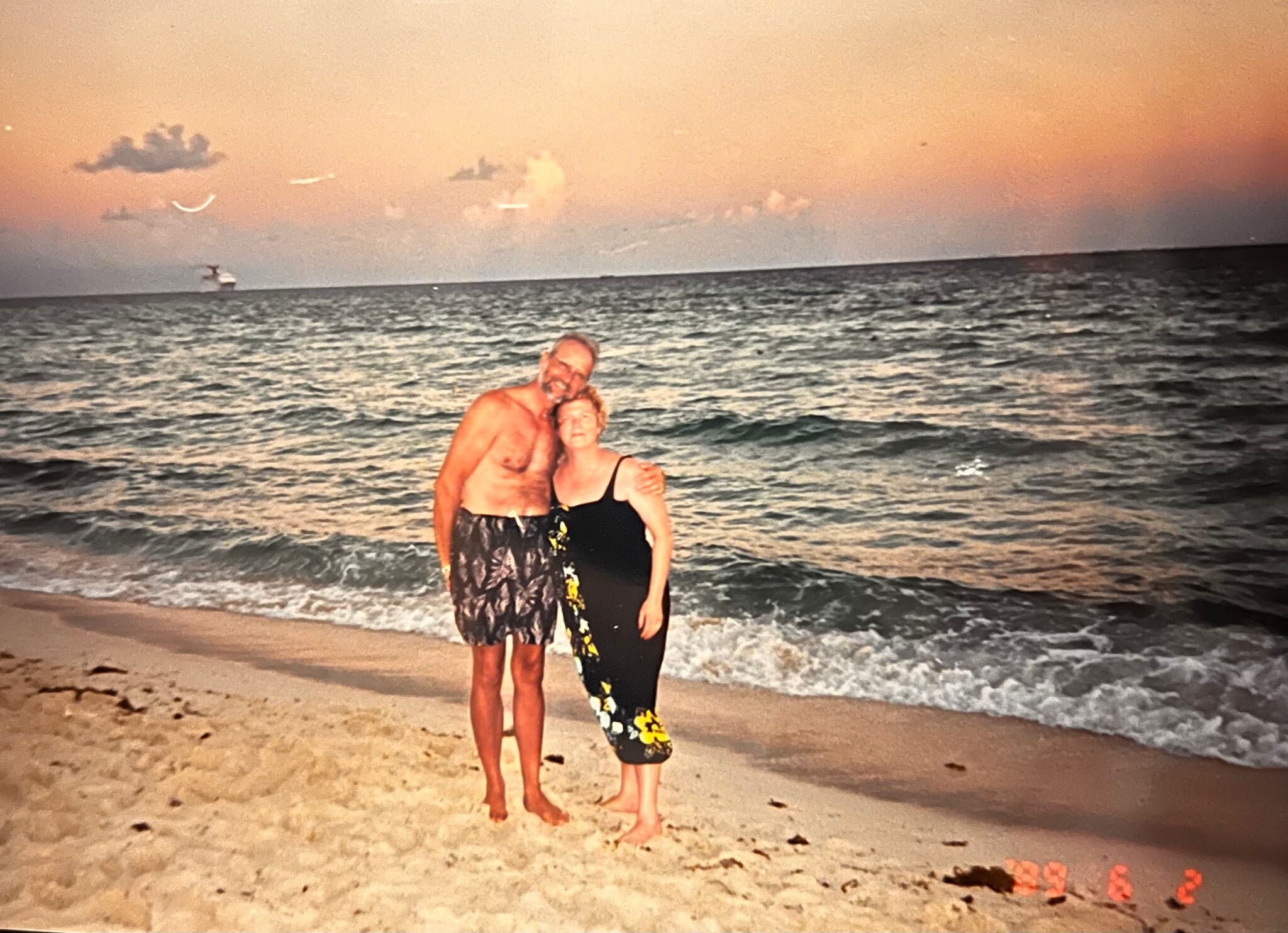 Two adults standing in shallow waves on a sandy beach at sunset, smiling and hugging.