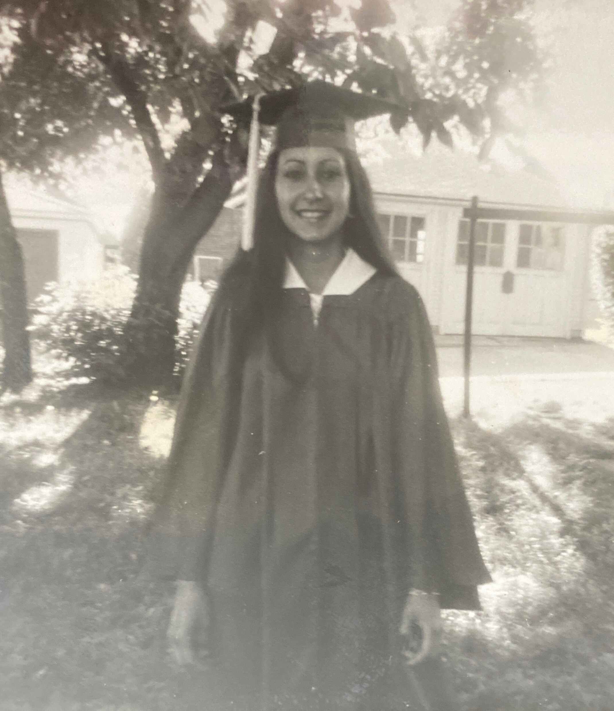 Young graduate in cap and gown smiling outdoors with a campus building in the background in black and white.