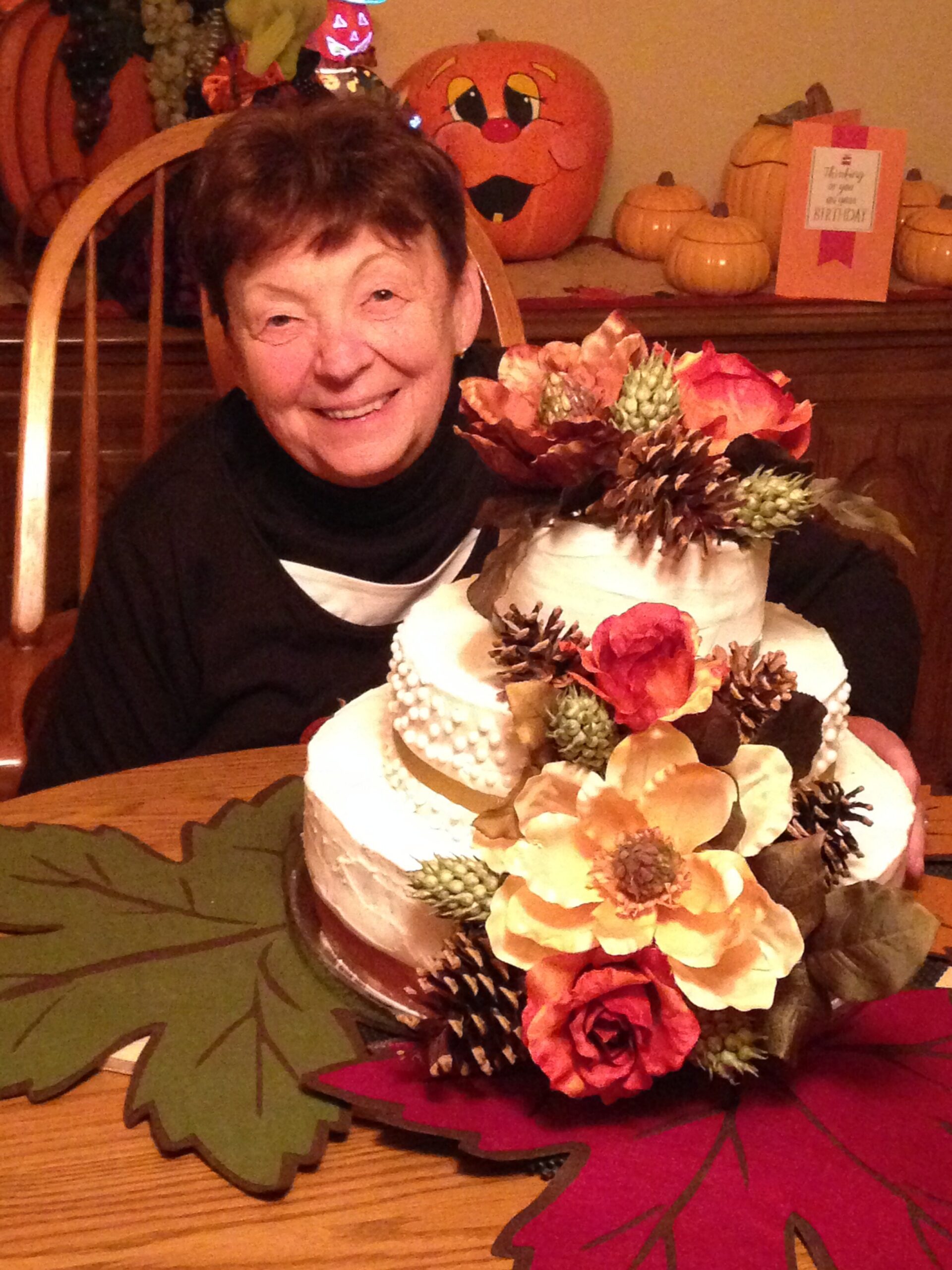 Smiling elderly woman with a layered autumn cake decorated with flowers, pinecones, and leaves; pumpkins in the background at a festive table.