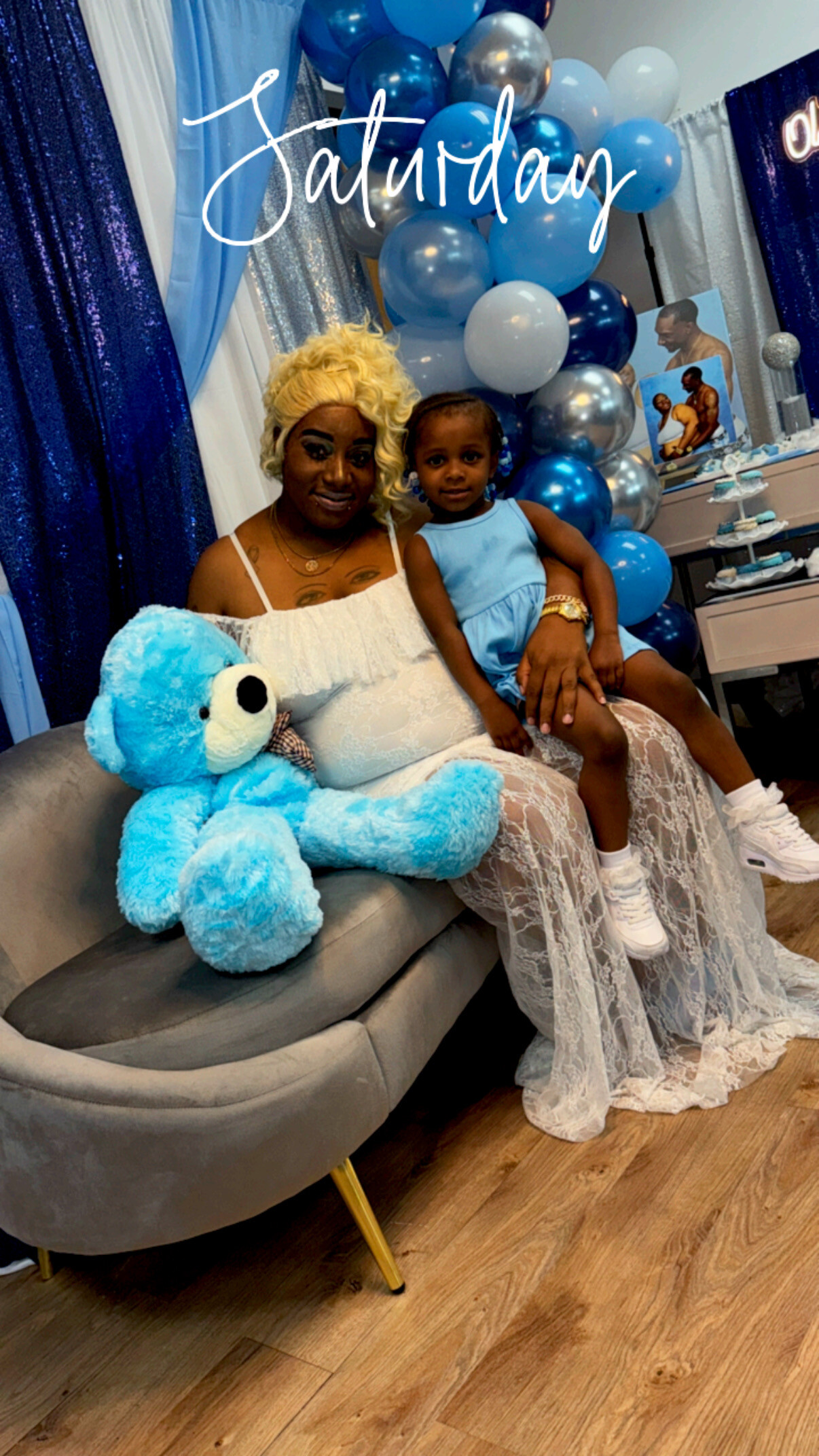 Woman with blonde hair and a young girl sit on a gray sofa at a blue-themed party, with a large blue teddy bear and balloons behind them; 'Saturday' text overlays the image.