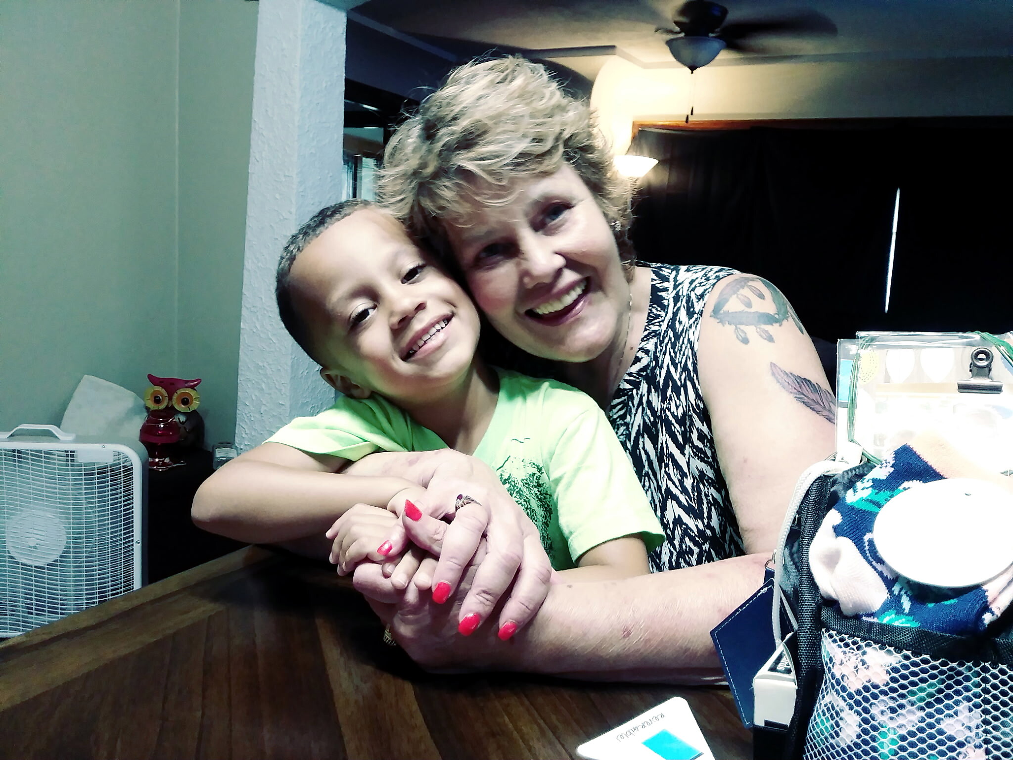 Smiling woman with short hair hugging a young child at a kitchen table indoors.