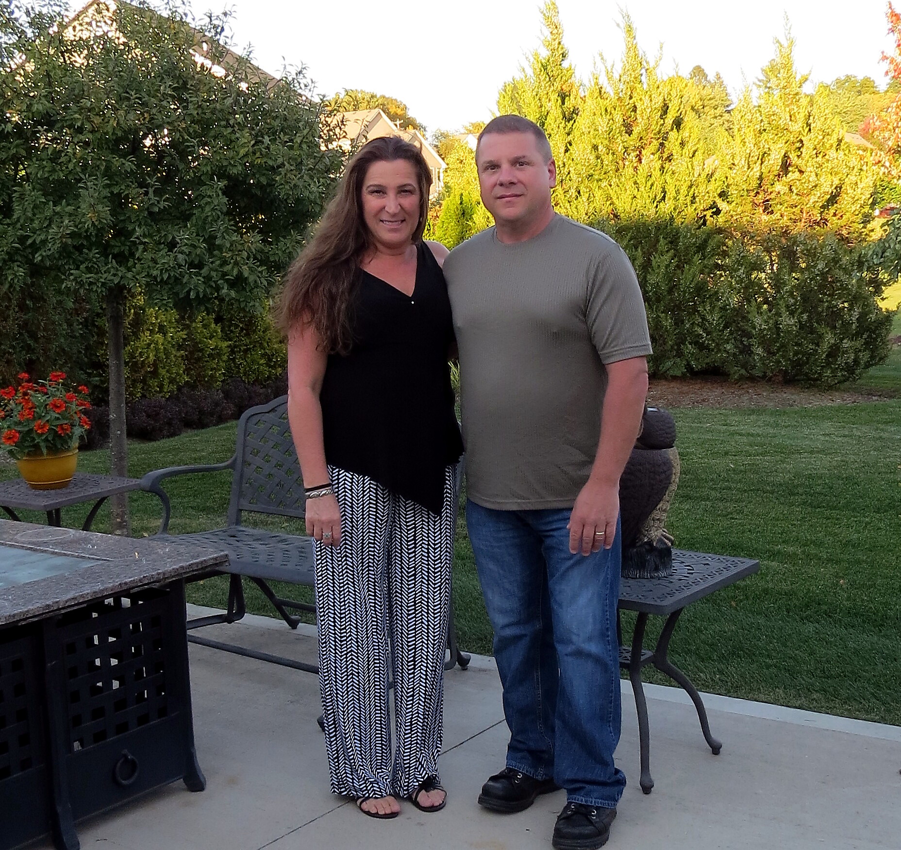A couple standing on a backyard patio, smiling at the camera with green hedges and trees behind them.