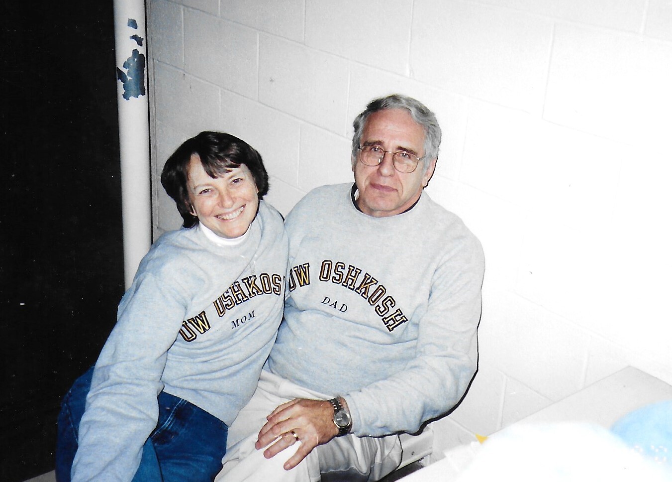 Smiling woman and man sit together, wearing matching gray sweatshirts that read 'UW Oshkosh' with 'Mom' and 'Dad' below the text.