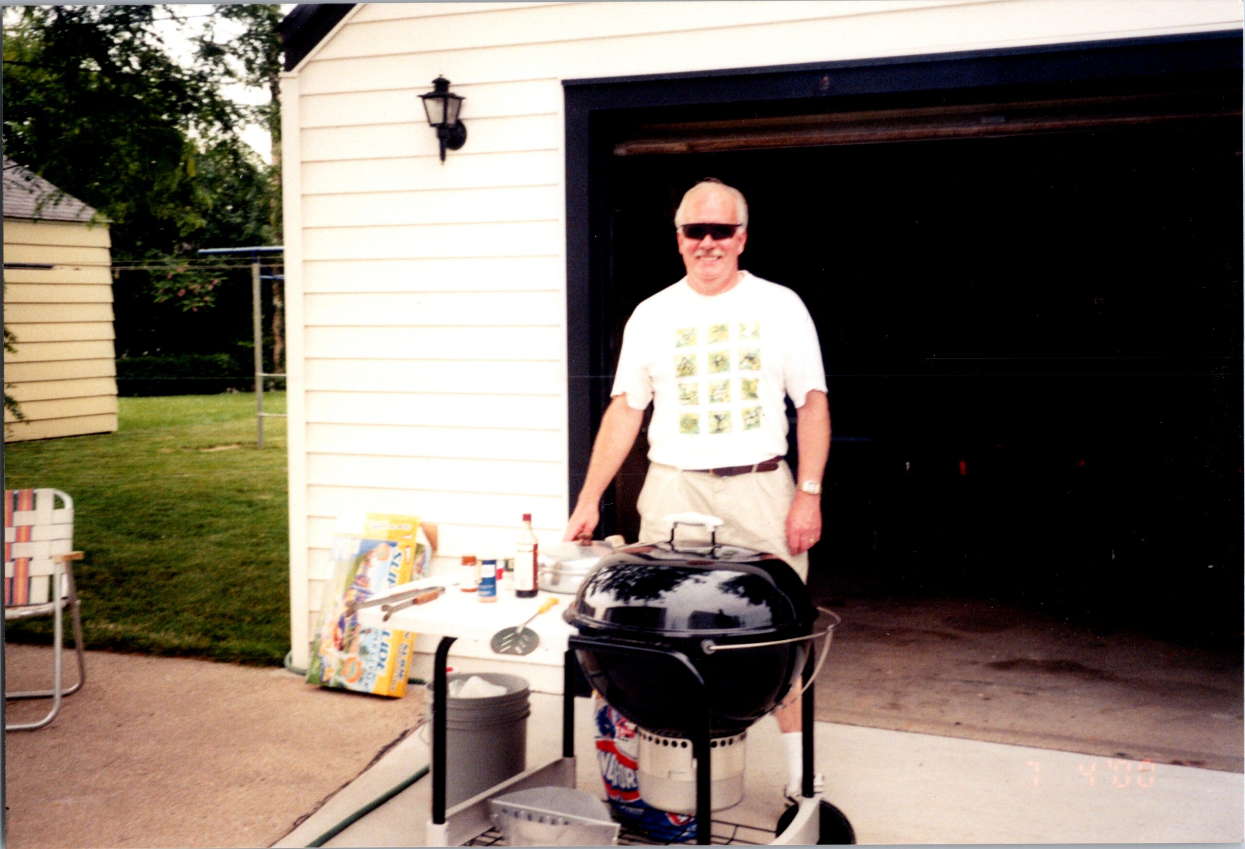 Older man in sunglasses near a barbecue grill on a patio by a cream-colored house.