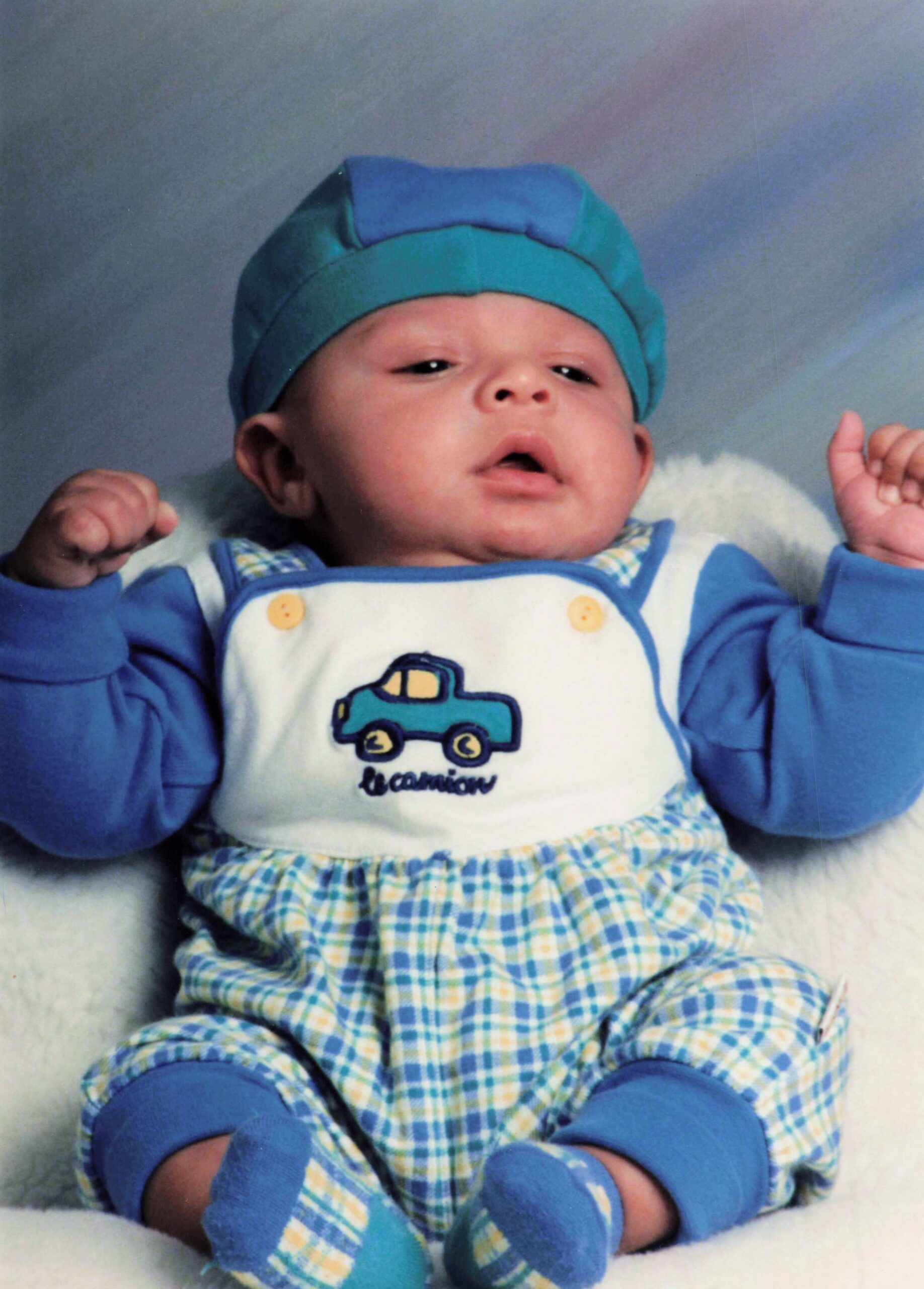 Infant wearing a blue hat and blue-and-plaid outfit with a small car illustration on the chest, lying on a light surface.