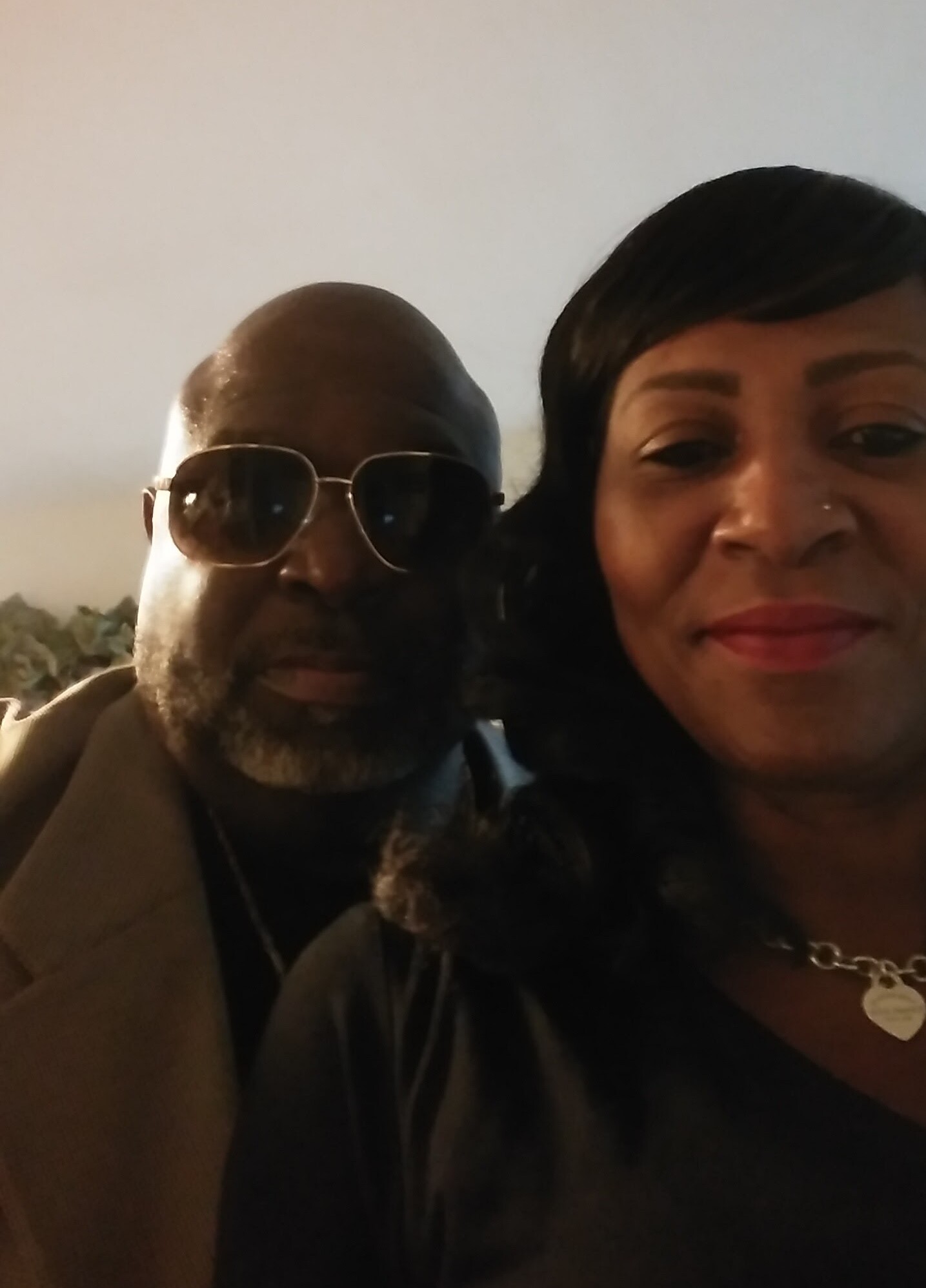 Close-up selfie of a man wearing sunglasses and a beard beside a woman with short dark hair, indoors.