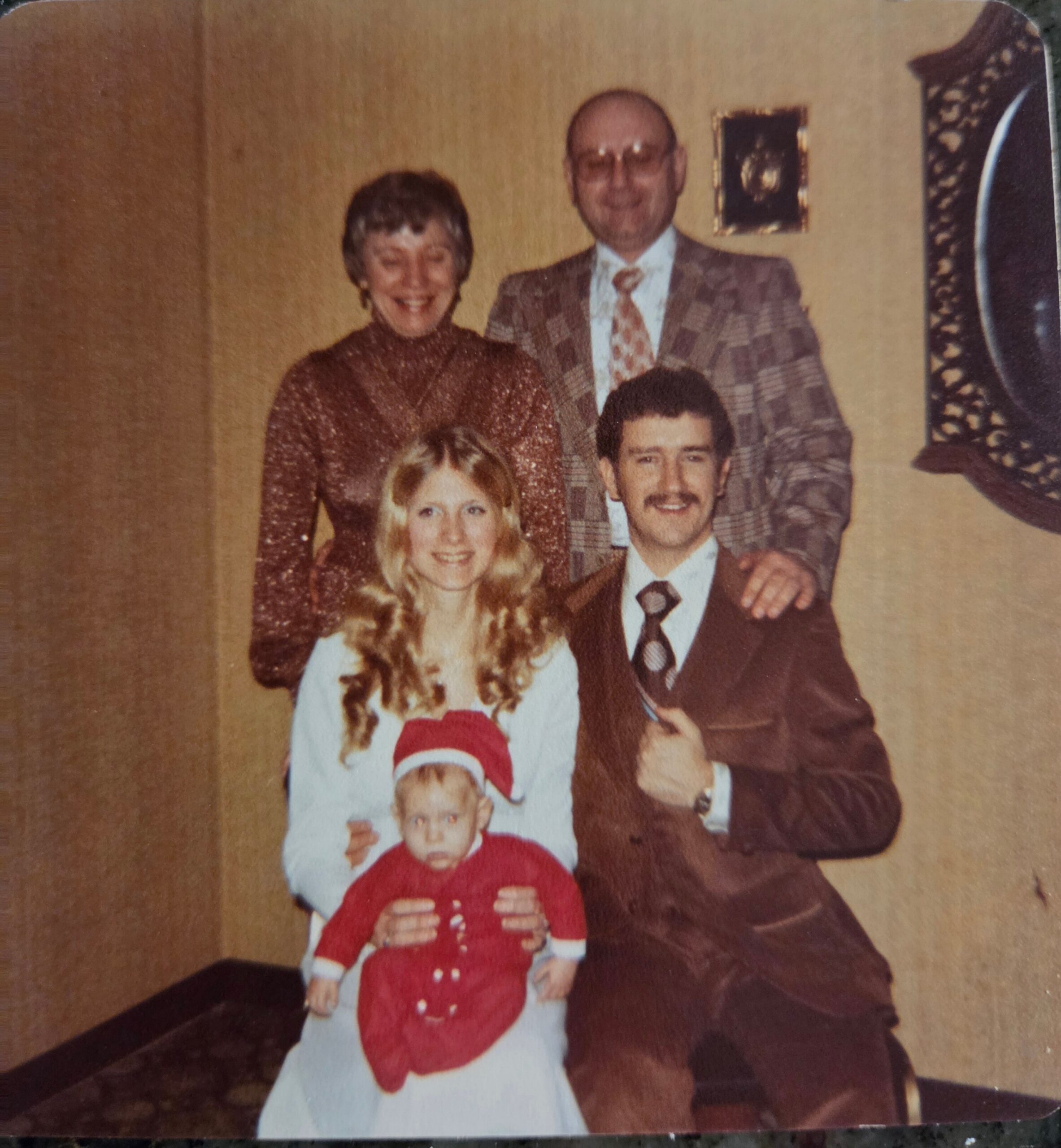 A vintage family portrait: two adults standing behind a smiling pair, with a baby in a red Santa outfit at the front.