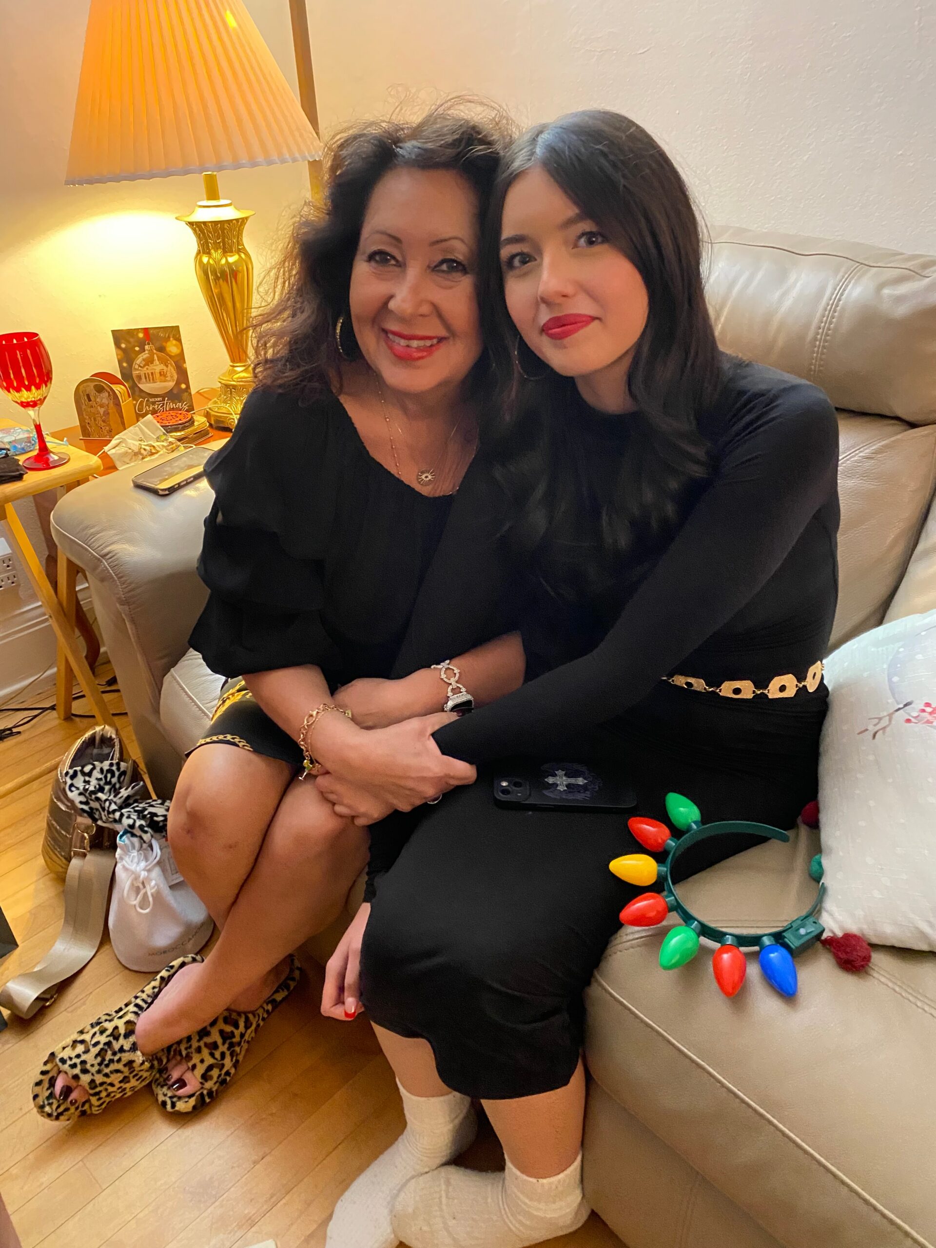 Two women sitting together on a beige sofa, smiling at the camera; one with curly dark hair, the other with straight dark hair, wearing black outfits and red lipstick, with Christmas lights nearby on the couch area.