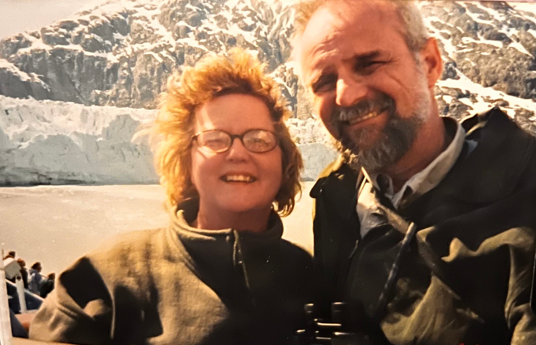 Two adults wearing winter jackets smile for a selfie in a snowy mountain landscape behind them, with rugged cliffs in view.
