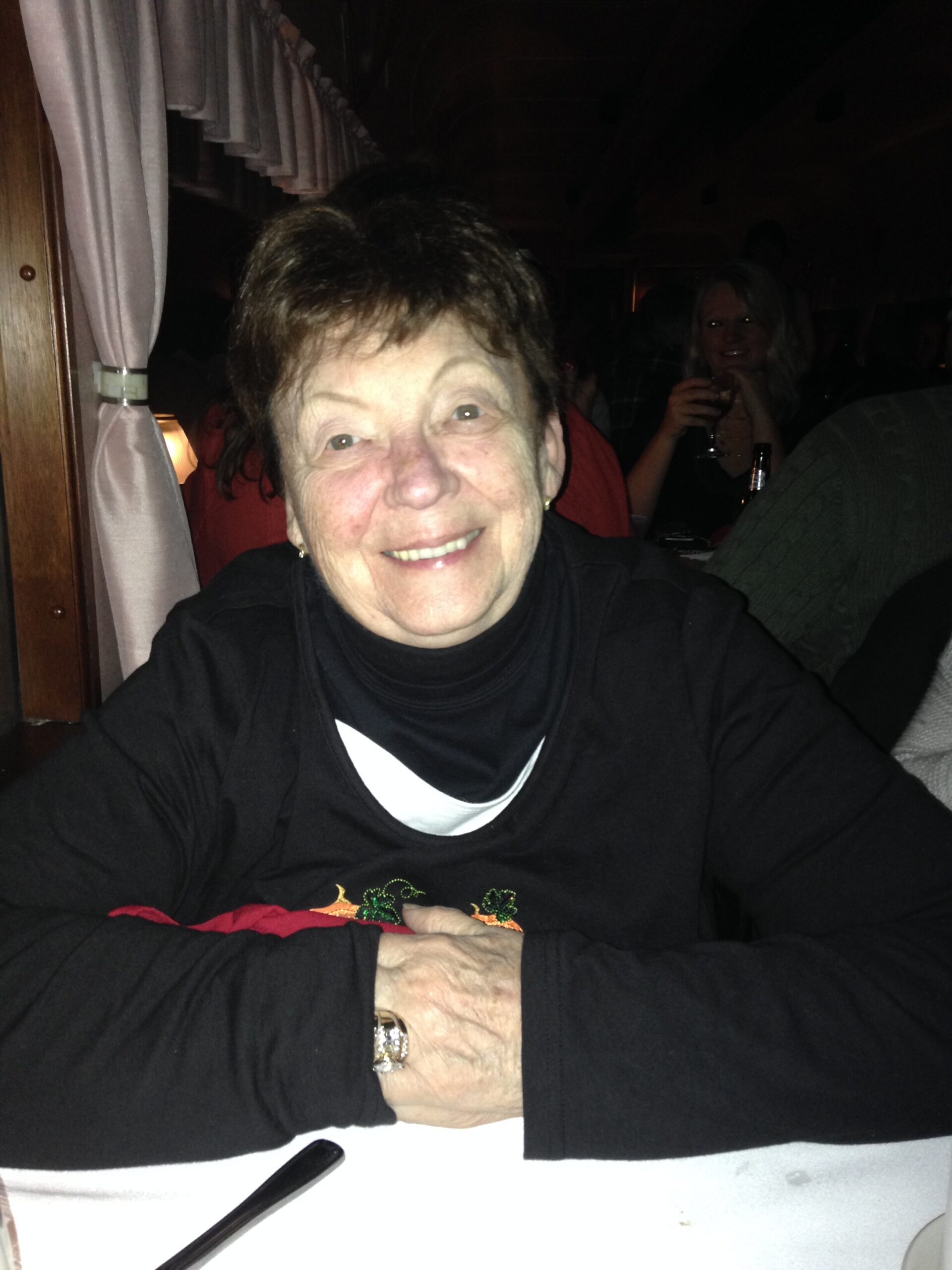 Smiling older woman with short hair in a black top, seated at a table indoors with a white surface in front of her.