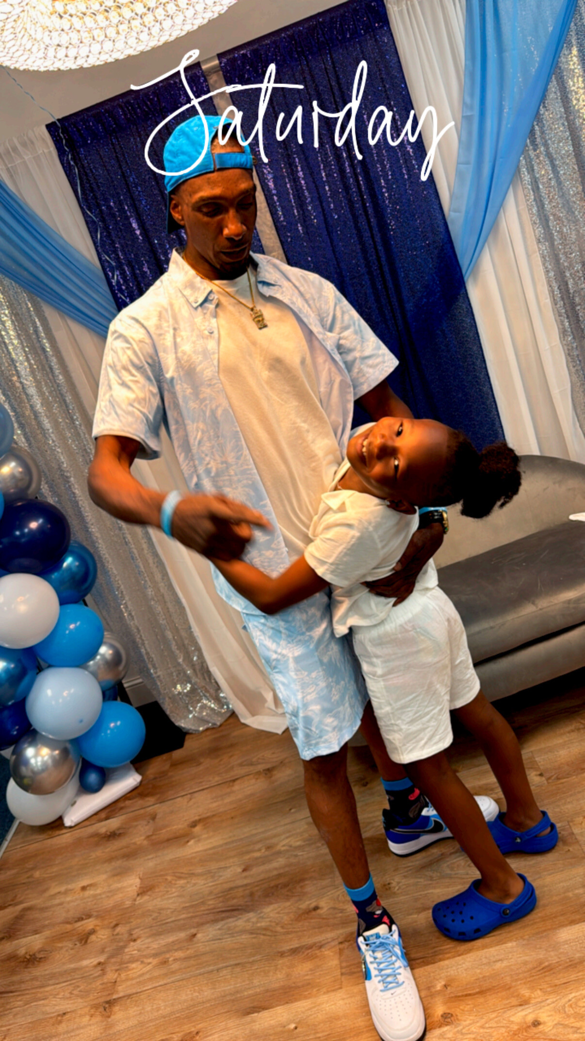 A man in a light blue shirt and backwards cap dancing with a smiling boy in white at a blue-themed party backdrop labeled 'Saturday'.