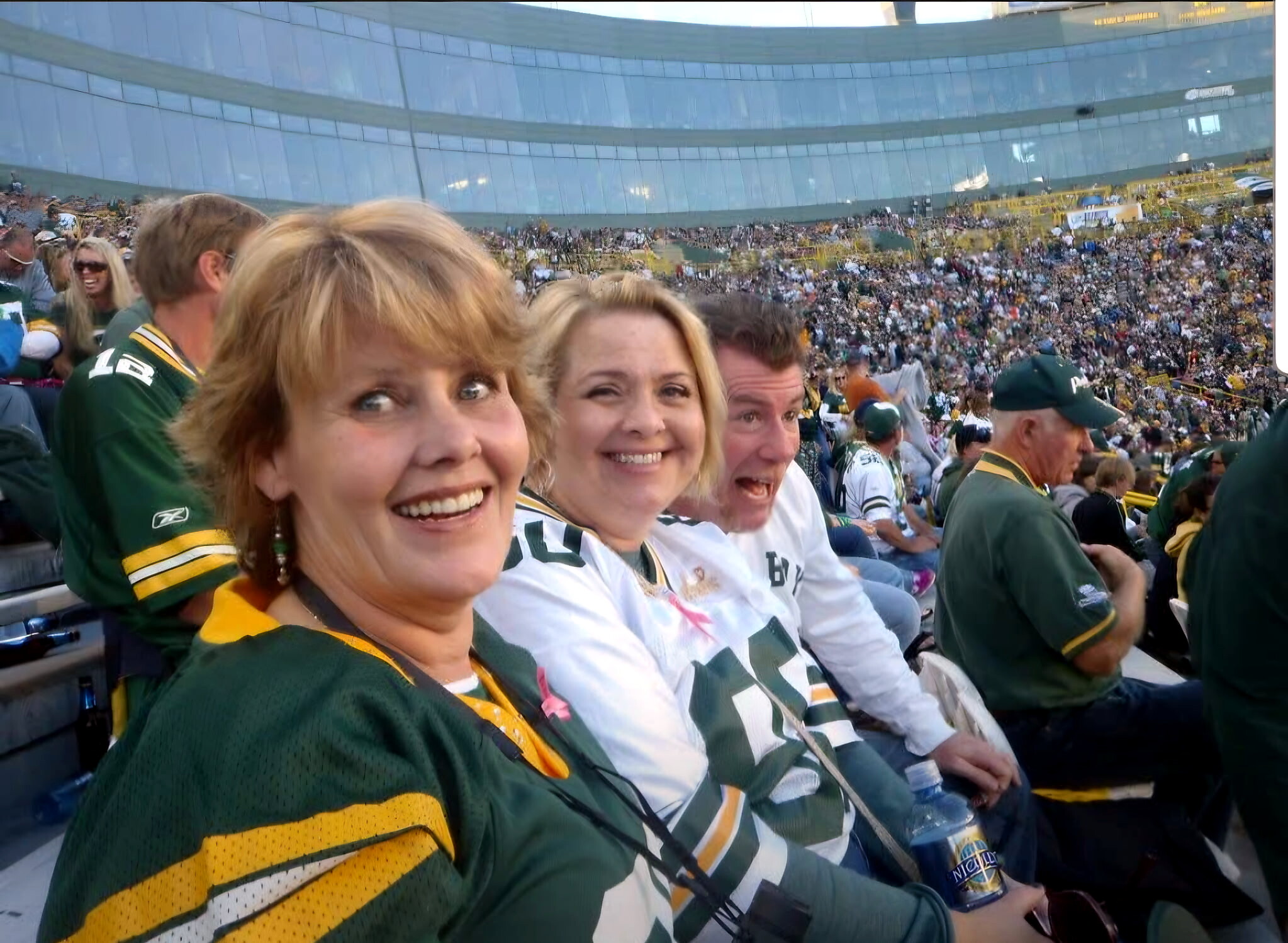 Smiling fans in green and gold jerseys take a selfie at a packed football stadium.