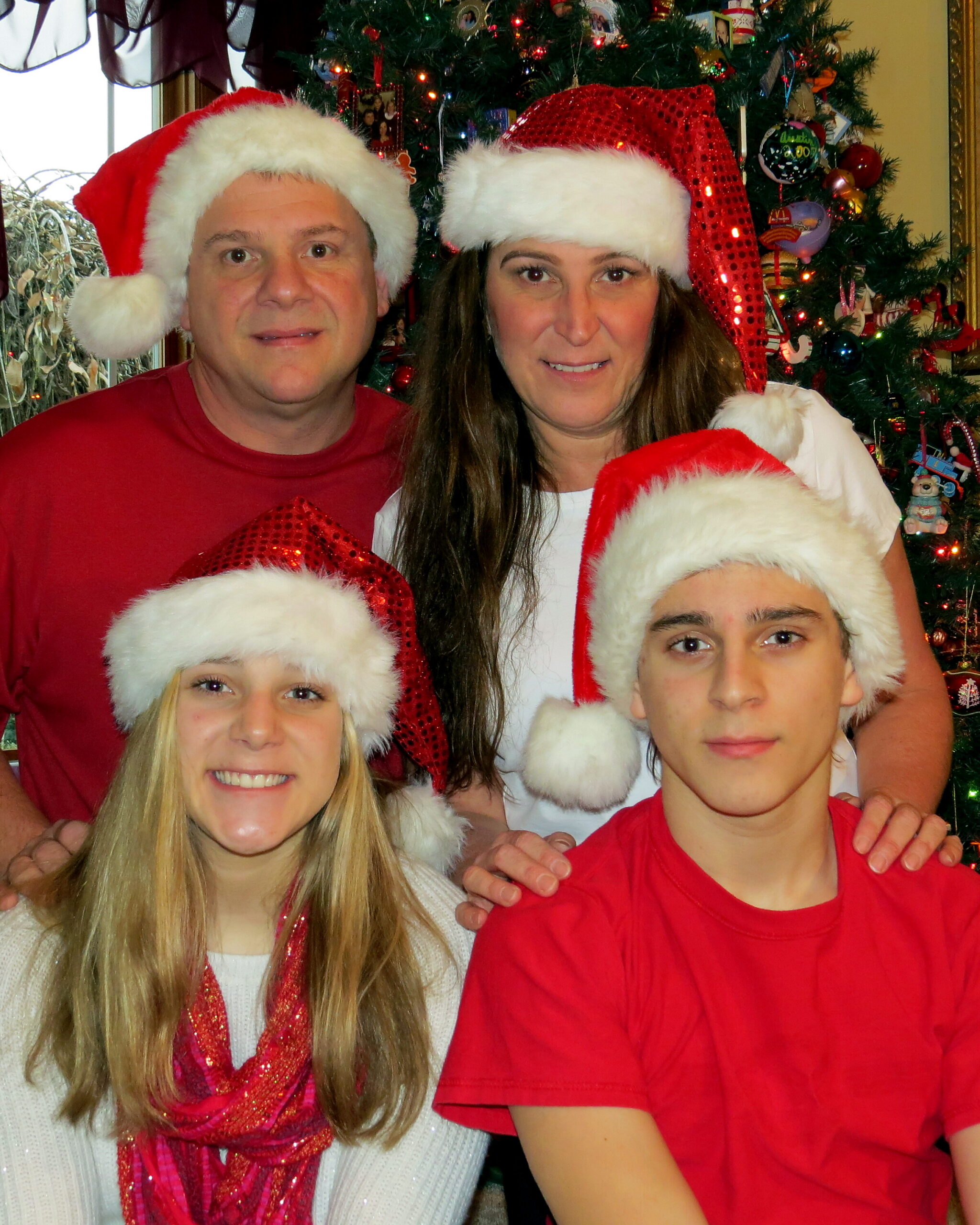 Four people wearing Santa hats posing in front of a decorated Christmas tree in a festive home setting.