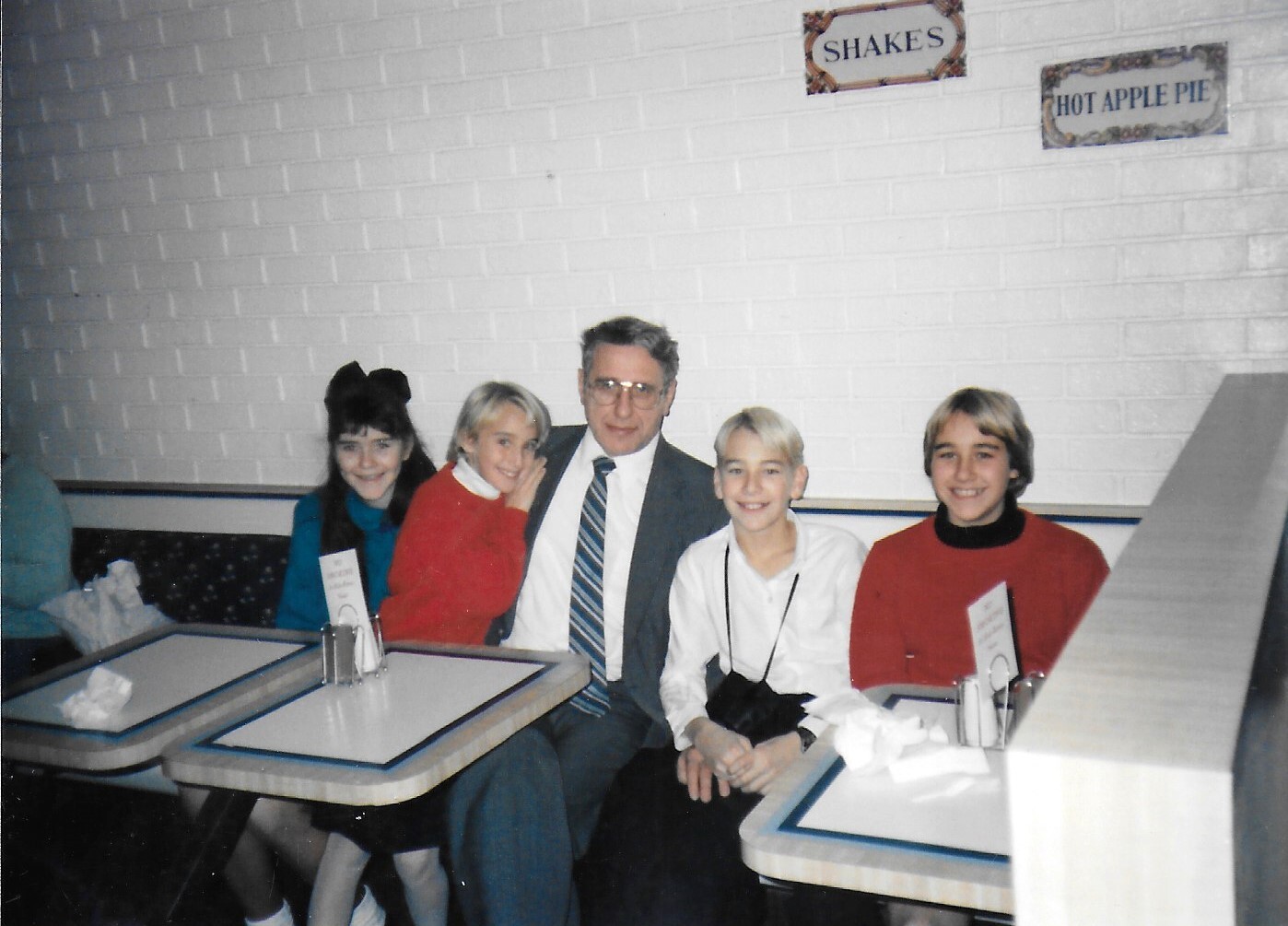 Man in suit and tie seated with four smiling kids in a retro diner booth.