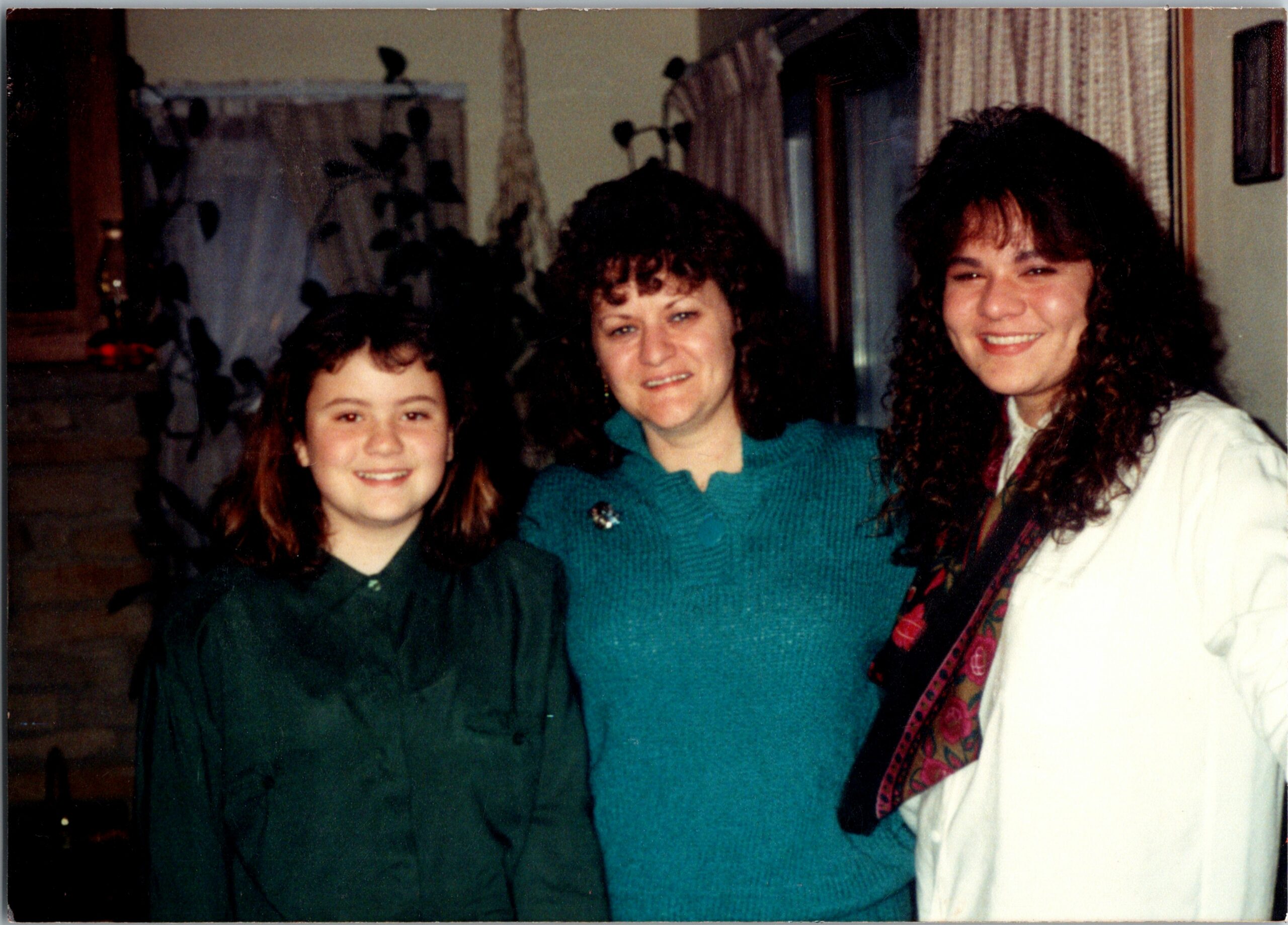 Three women posing indoors, smiling: a girl in a dark green dress on the left, a woman in a teal sweater in the middle, and a woman in white with a patterned scarf on the right.