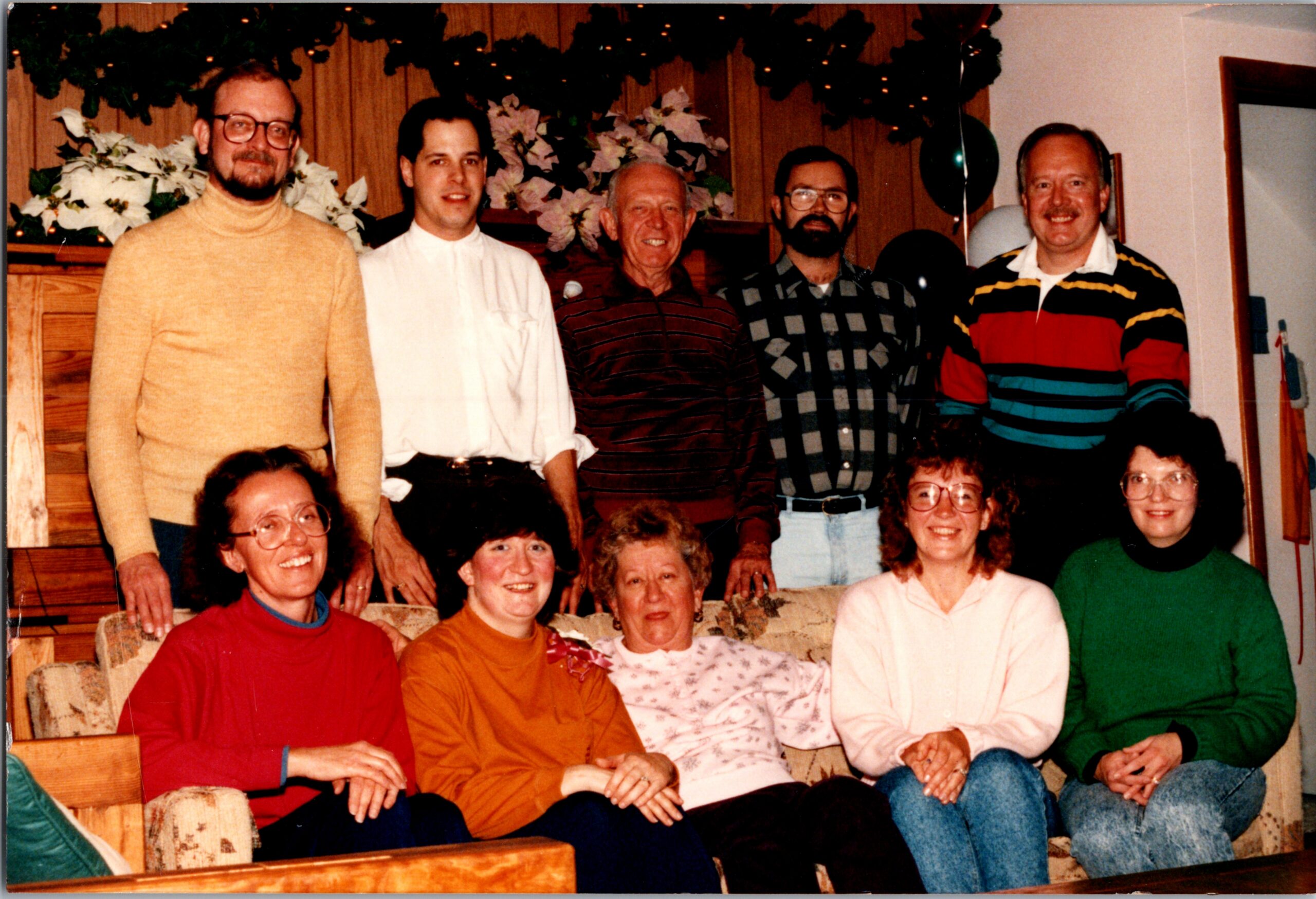 A group of adults posing for a casual holiday photo indoors, wearing colorful sweaters and smiling on a sofa and standing behind it.
