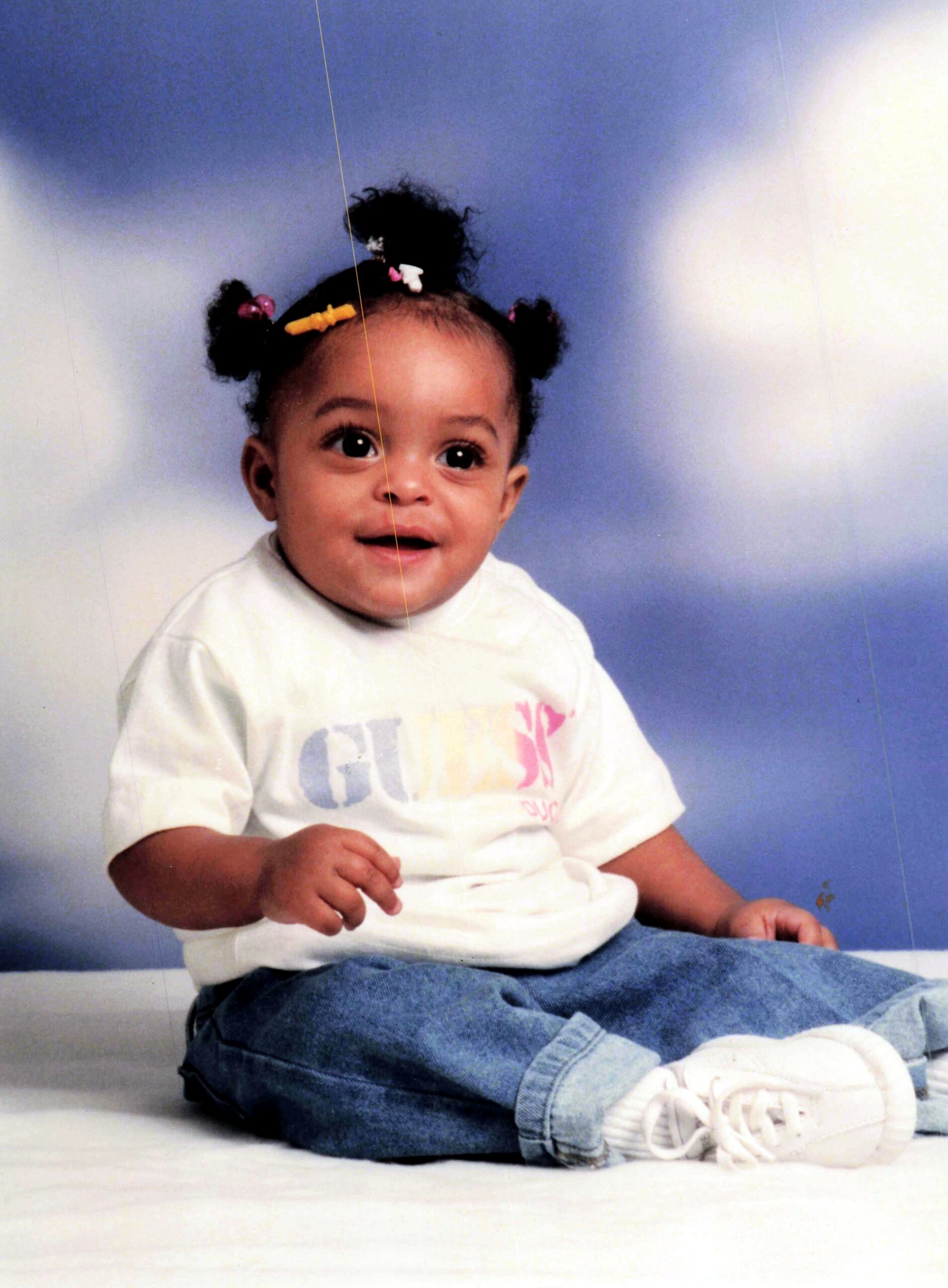 Toddler seated on a white backdrop, wearing a white shirt, blue jeans, and white sneakers, smiling at the camera with puffy hair