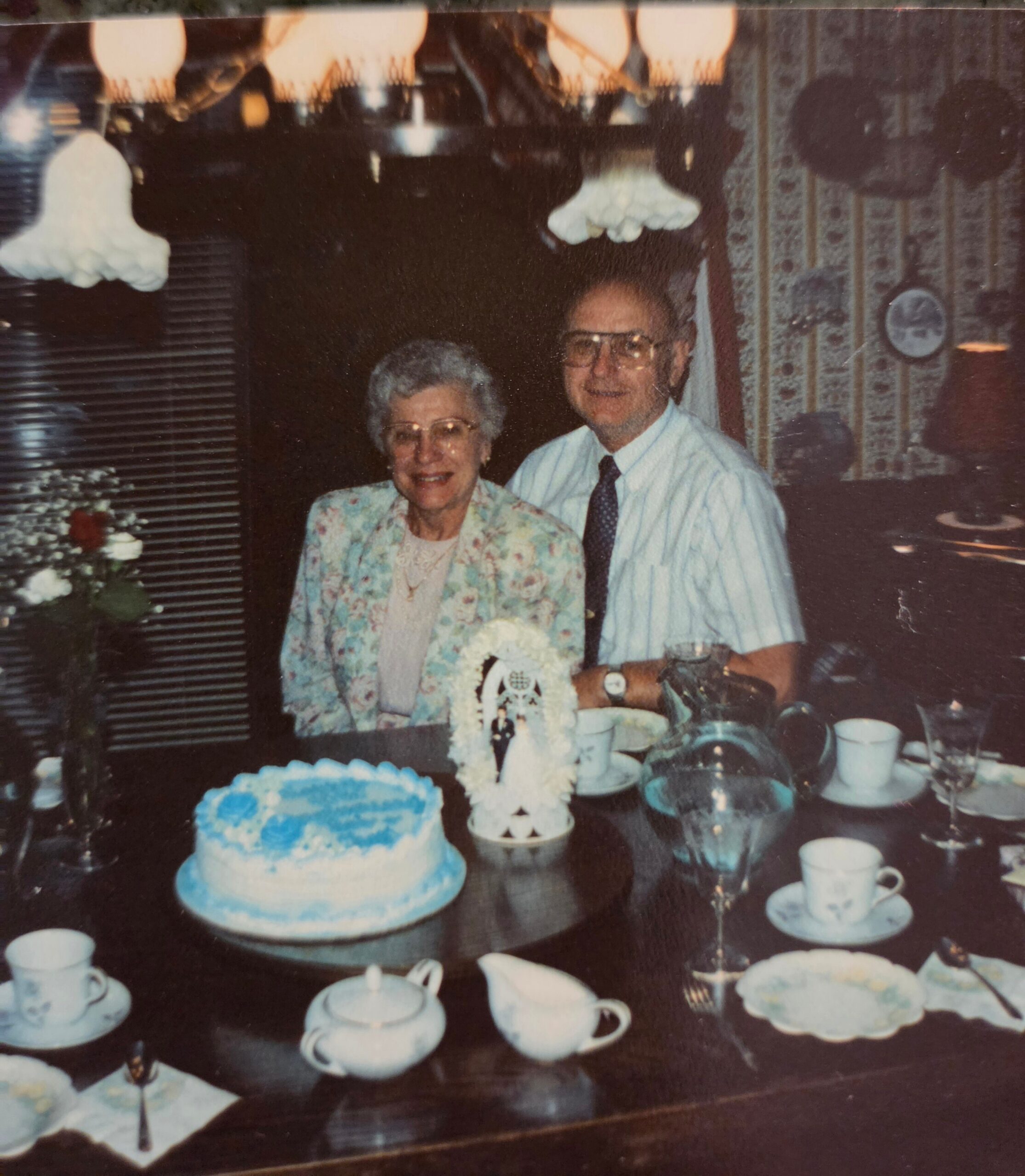 Elderly couple smiling at a birthday celebration with a blue frosted cake on the table in front of them.