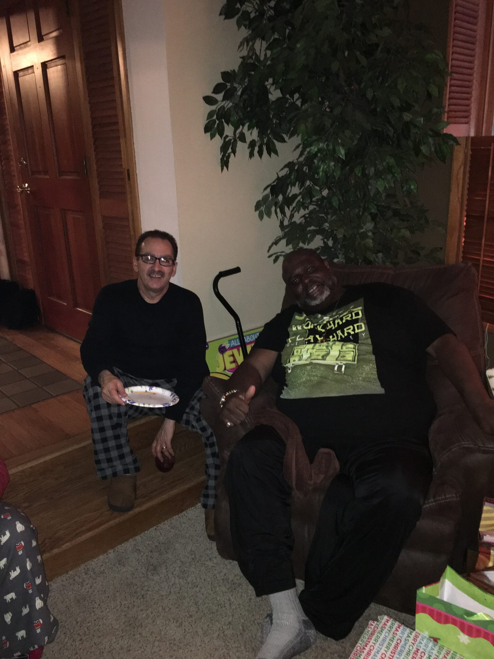 Two men sit in a living room; one white man with glasses holds a plate, the other Black man leans back on a couch, both smiling, with a large plant and gift bags in the background.