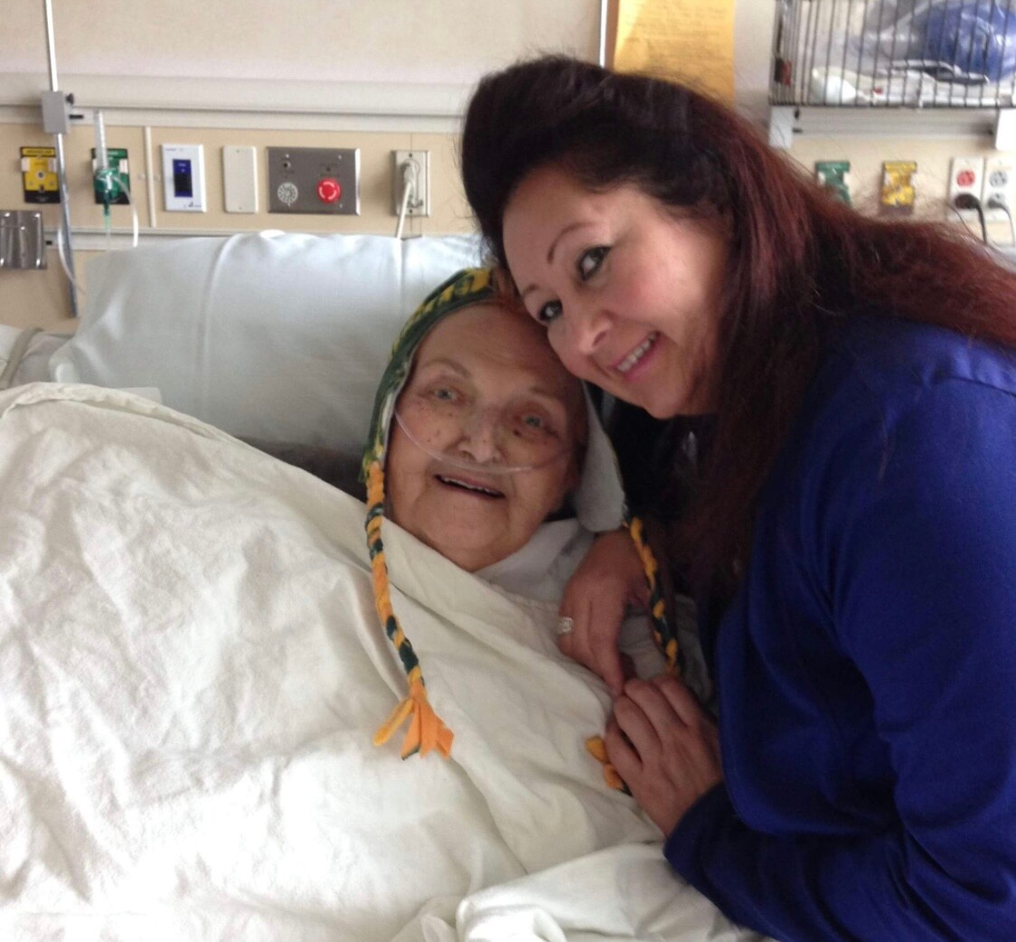 Elderly patient in a hospital bed with an oxygen tube, wearing a colorful knit hat and blanket, smiling as a younger woman in blue leans in for a hug.