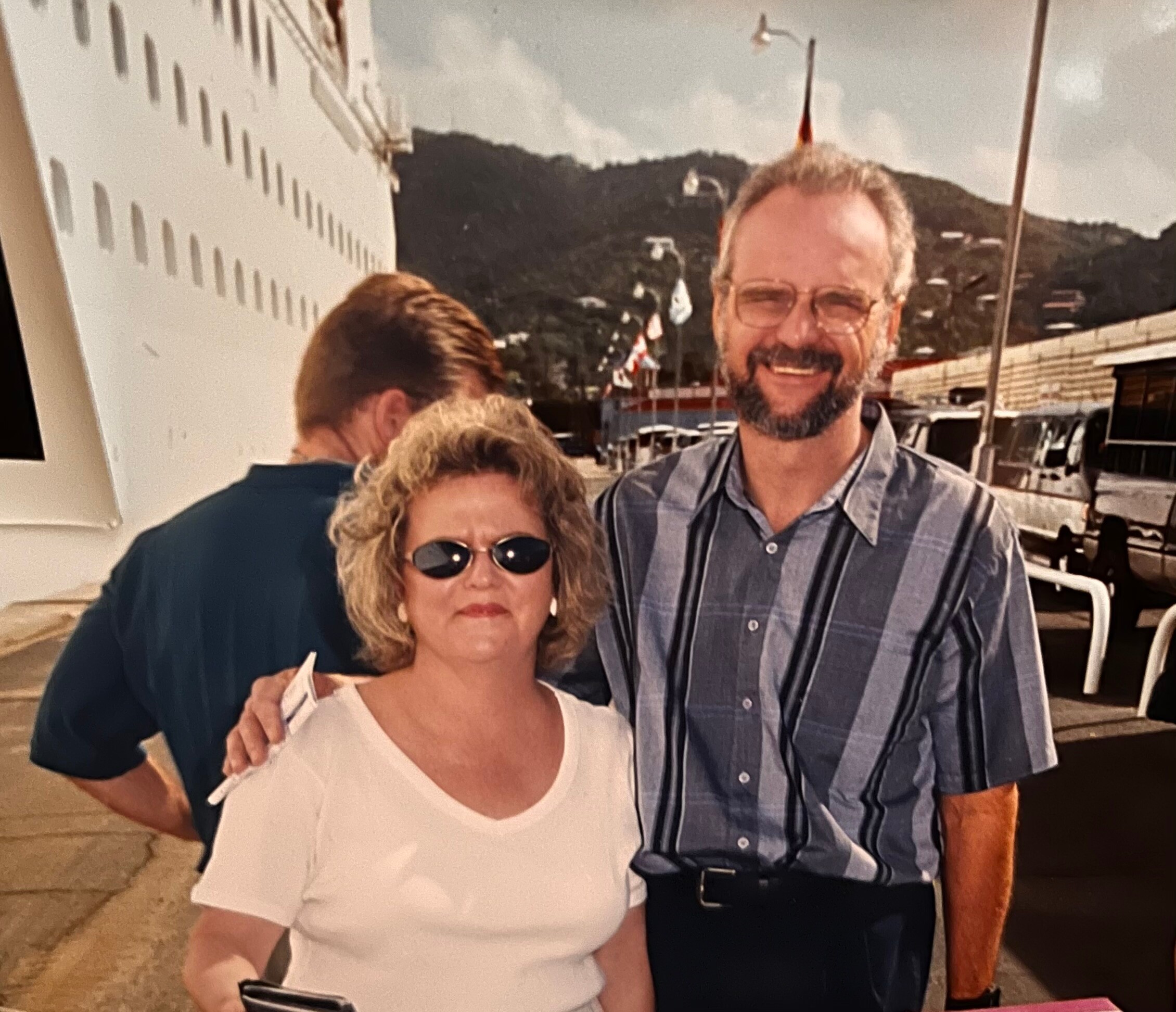 Smiling couple posing together outdoors, the man in a striped shirt and the woman in a white top with sunglasses.