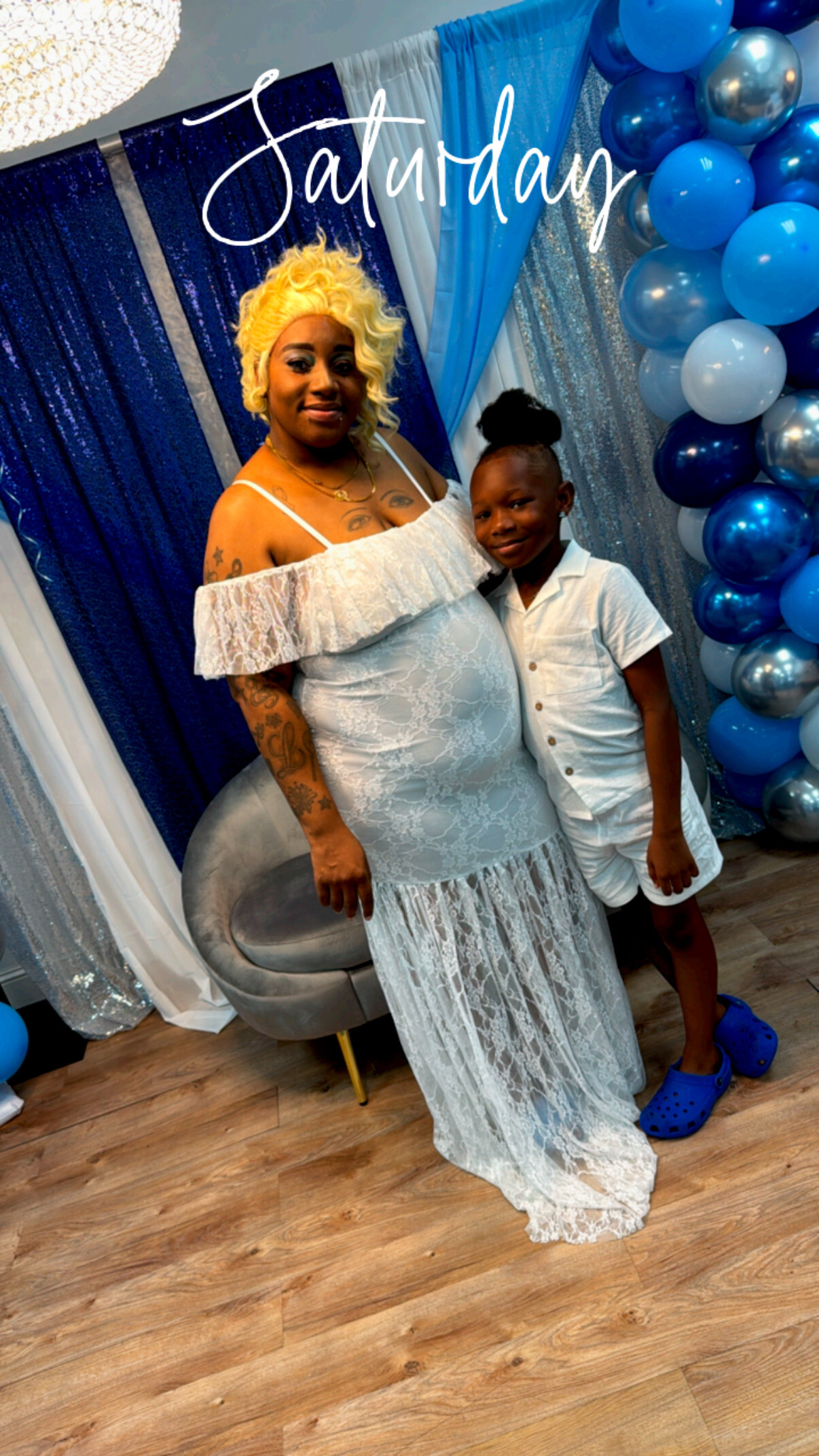 Woman in a light blue off-shoulder lace dress with a blonde wig stands beside a smiling child in white, at a blue-themed backdrop with balloons labeled Saturday.